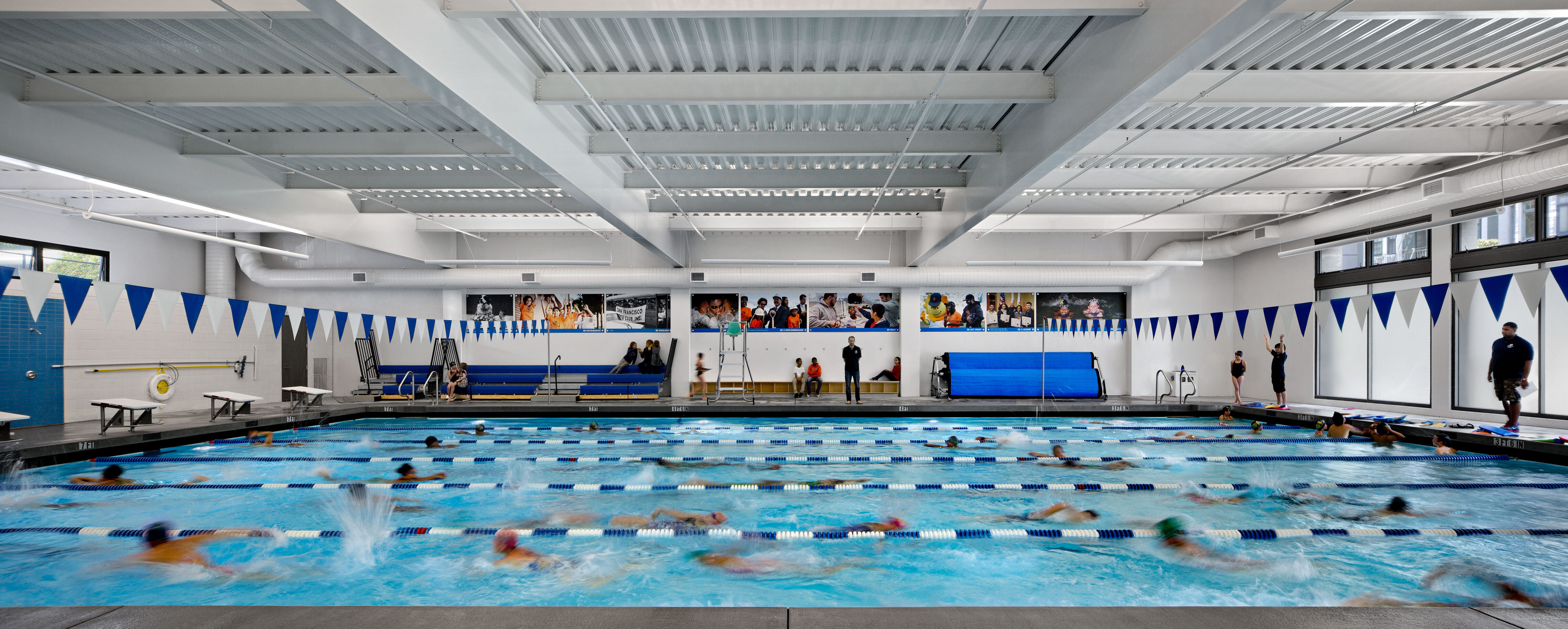 Wide view of an indoor swimming pool filled with children practicing laps in marked lanes. Blue and white triangular flags hang overhead, with spectators and coaches watching from the bleachers along the wall. The bright space features high ceilings, large windows, and colorful wall graphics celebrating teamwork and activity.