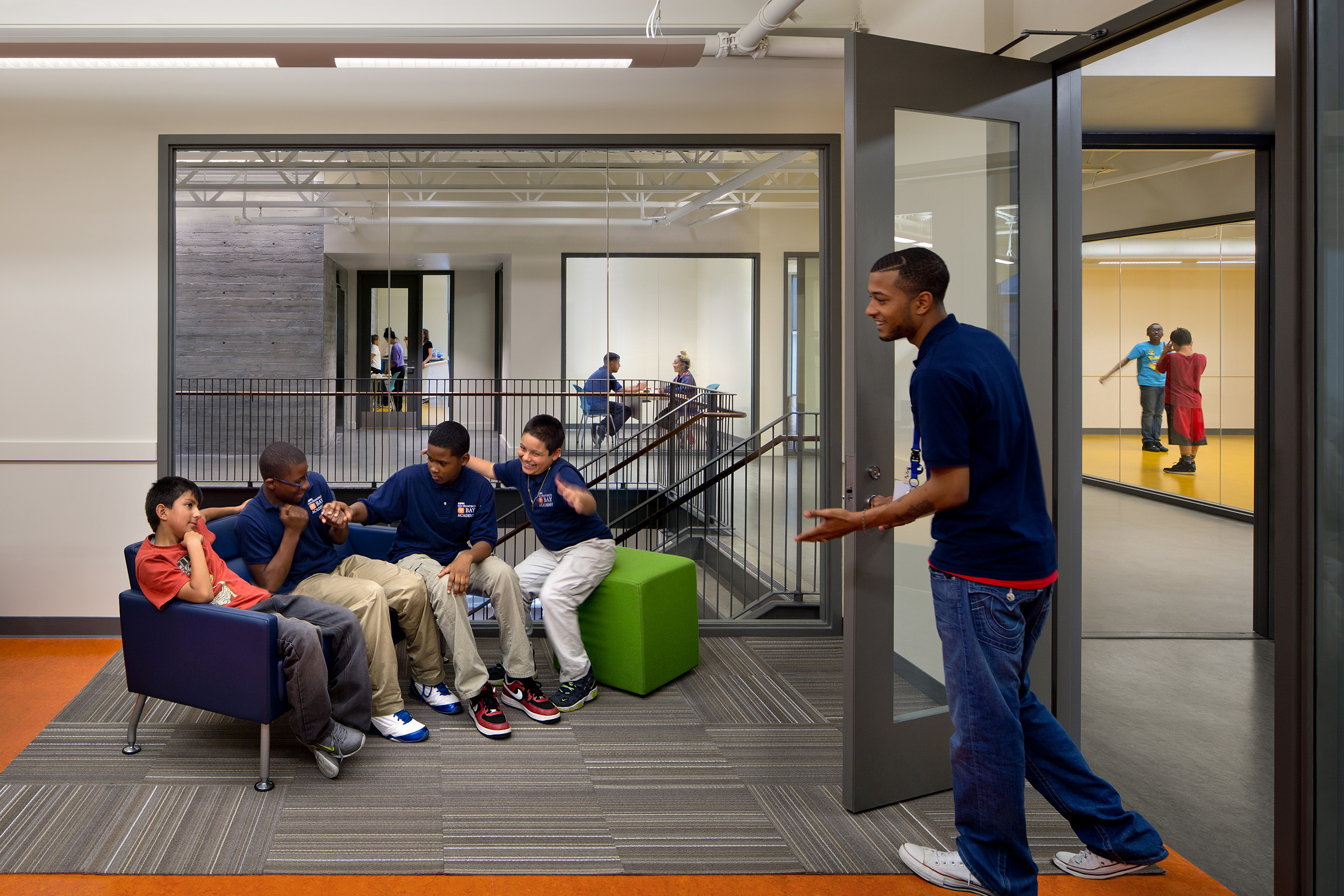 A youth mentor engages with a group of boys sitting and chatting in a common area. The open interior shows connected rooms and a dance studio beyond, creating an active, social atmosphere.