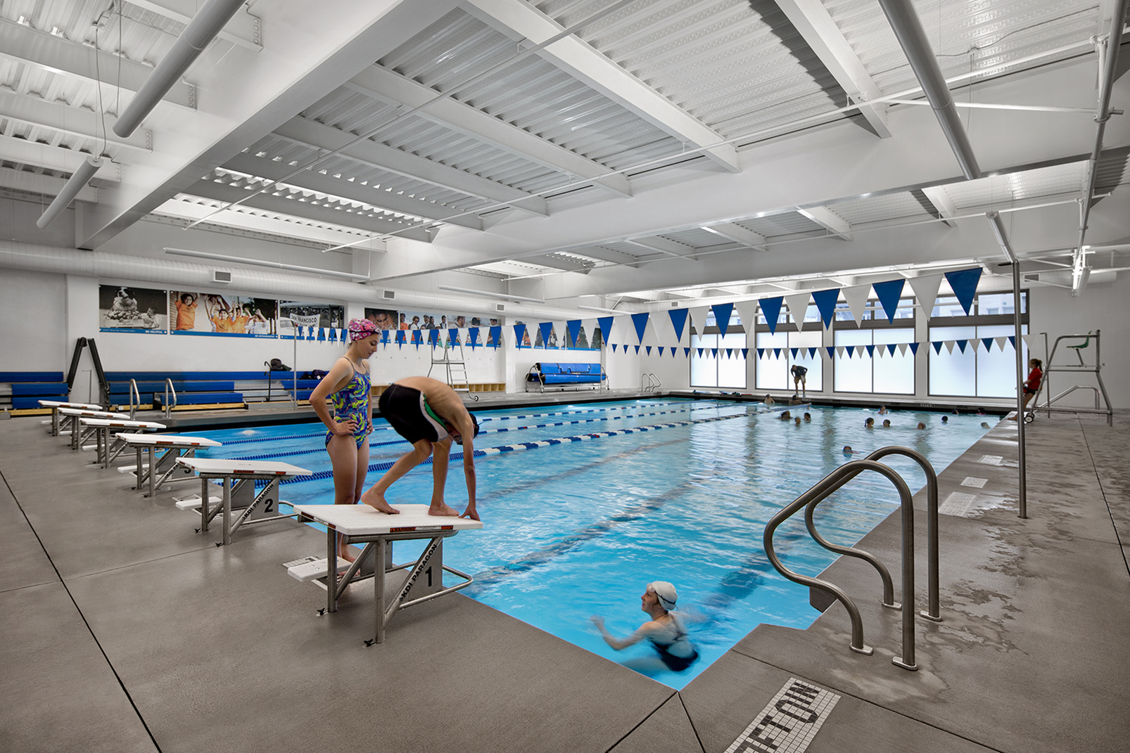 Swimmers practice in a bright indoor swimming pool lined with blue-and-white lane dividers and matching triangular flags overhead. Natural light filters through large frosted windows, and young swimmers stand on starting blocks preparing to dive.