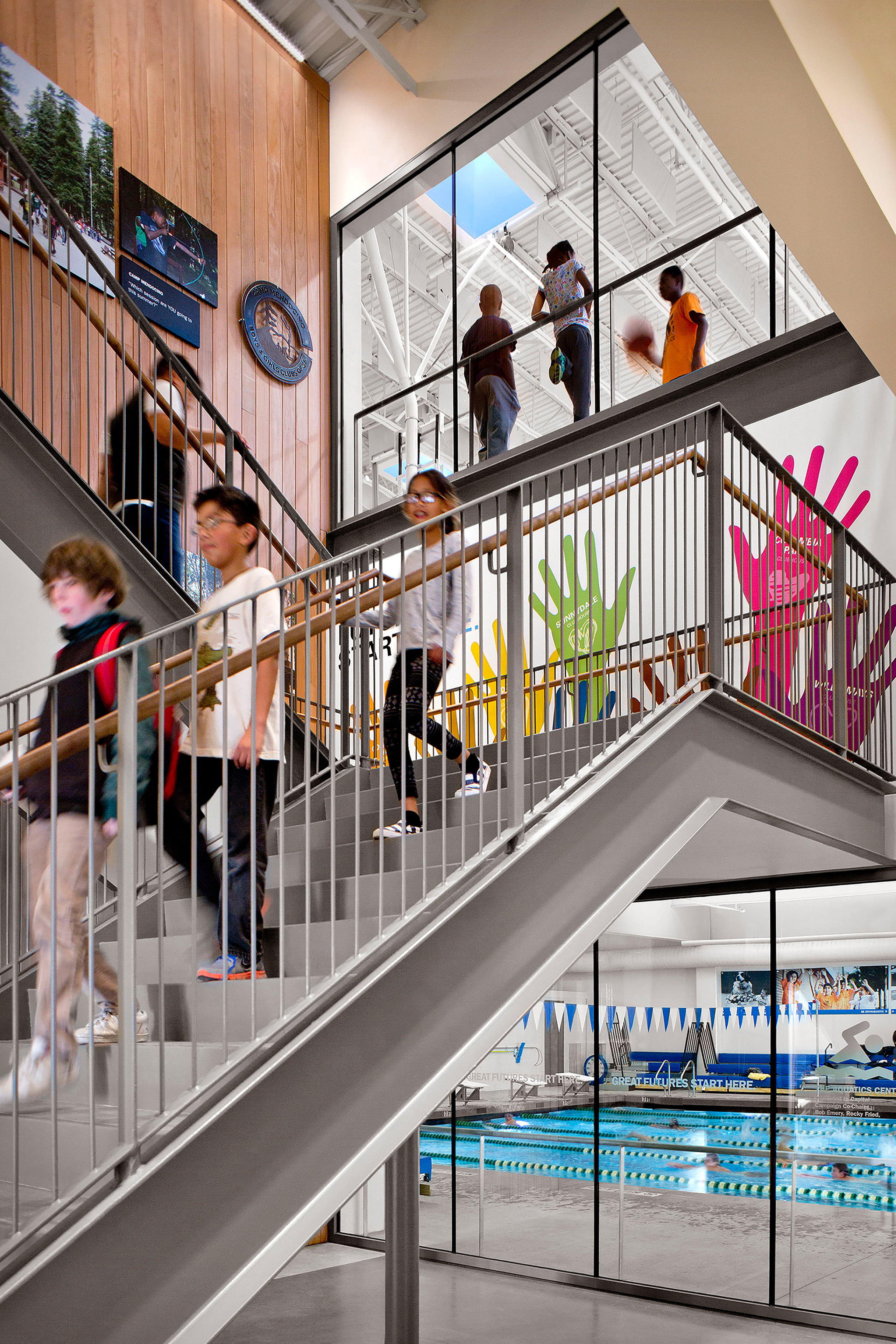 Students walk up and down a modern indoor stairwell in a bright community clubhouse building. Large windows reveal an indoor swimming pool below and a gym above where people are playing basketball. The space features colorful handprint graphics on the wall and wood panel accents, creating a vibrant, active atmosphere.