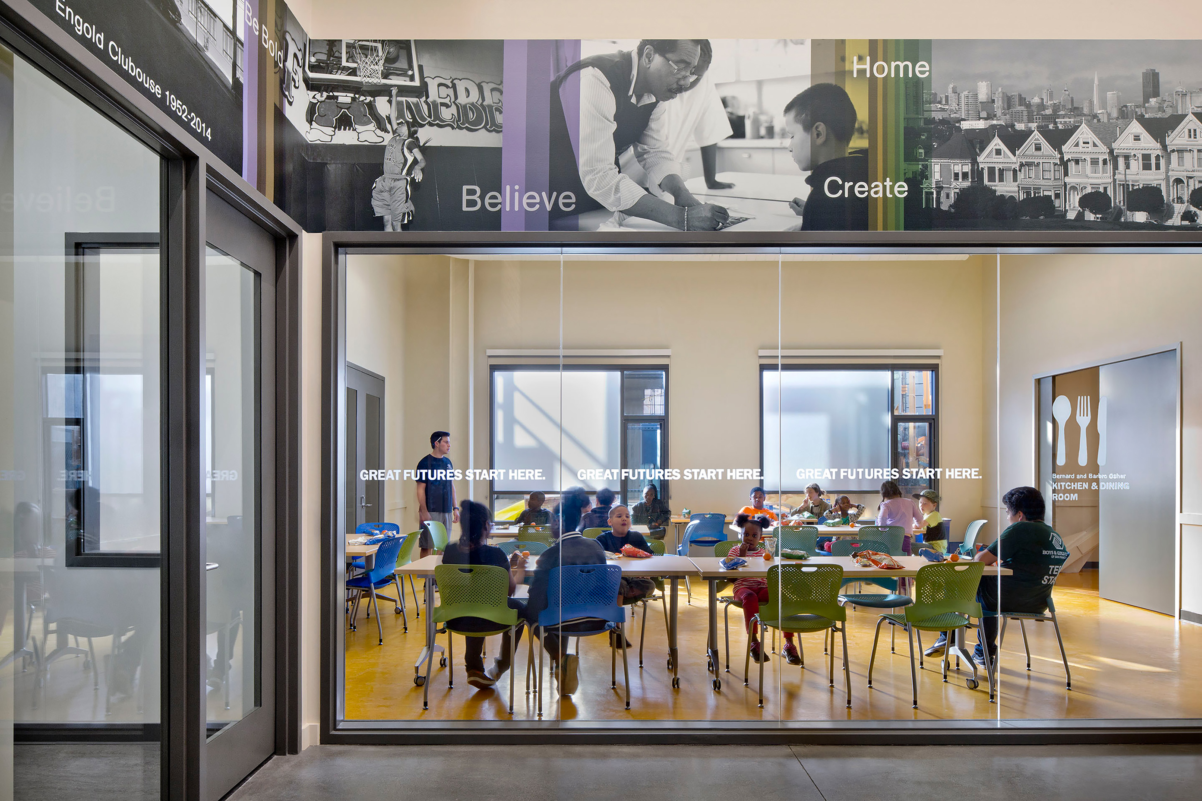 Children sit at colorful tables eating and socializing in a light-filled multipurpose dining room. A mural above the glass wall reads “Believe, Home, Create,” and the slogan “Great Futures Start Here” is printed across the glass panels.