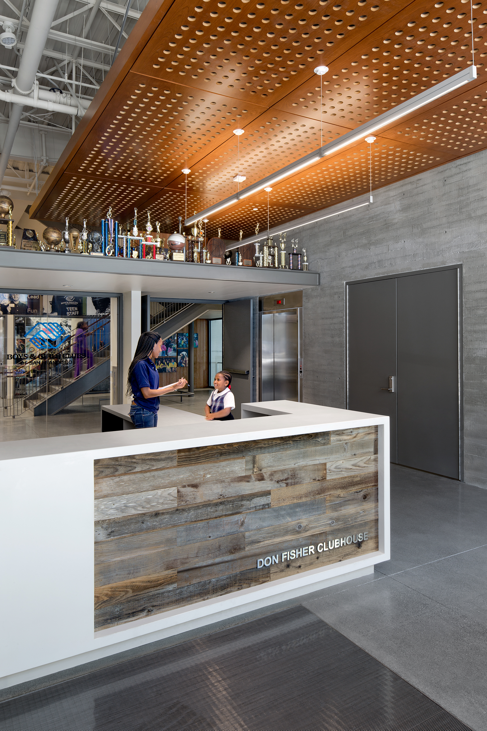 A reception area inside the Don Fisher Clubhouse featuring a modern white desk with reclaimed wood paneling. A staff member talks to a young child near the counter. Above, a perforated wood ceiling and a row of trophies add warmth and character to the bright, industrial-style interior.