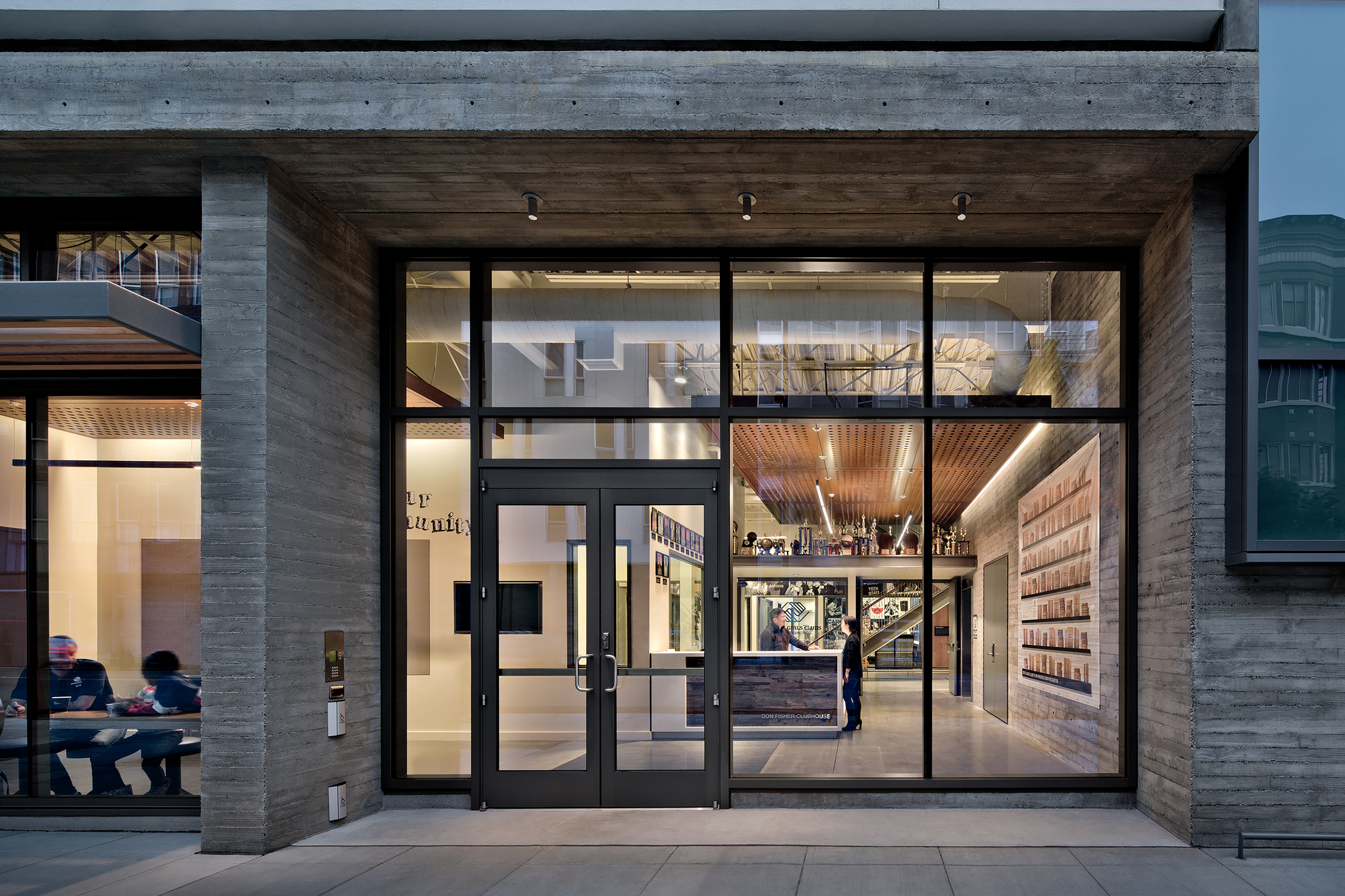 Exterior view of the Don Fisher Clubhouse entrance with floor-to-ceiling glass walls revealing the bright interior lobby. The reception desk, trophies, and wood-paneled ceiling are visible through the glass façade framed by textured concrete.