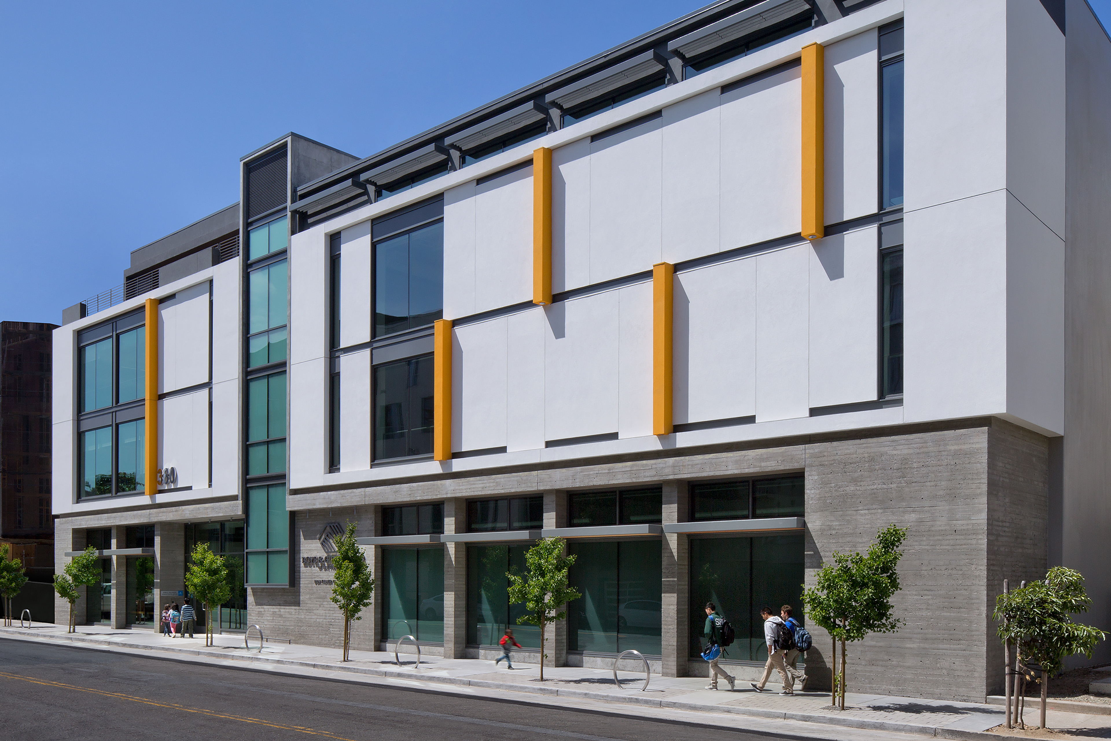 Street view of the Don Fisher Clubhouse building, featuring a modern white façade with tall windows and bold yellow vertical fins. Pedestrians walk along the sidewalk lined with young trees under a bright blue sky.