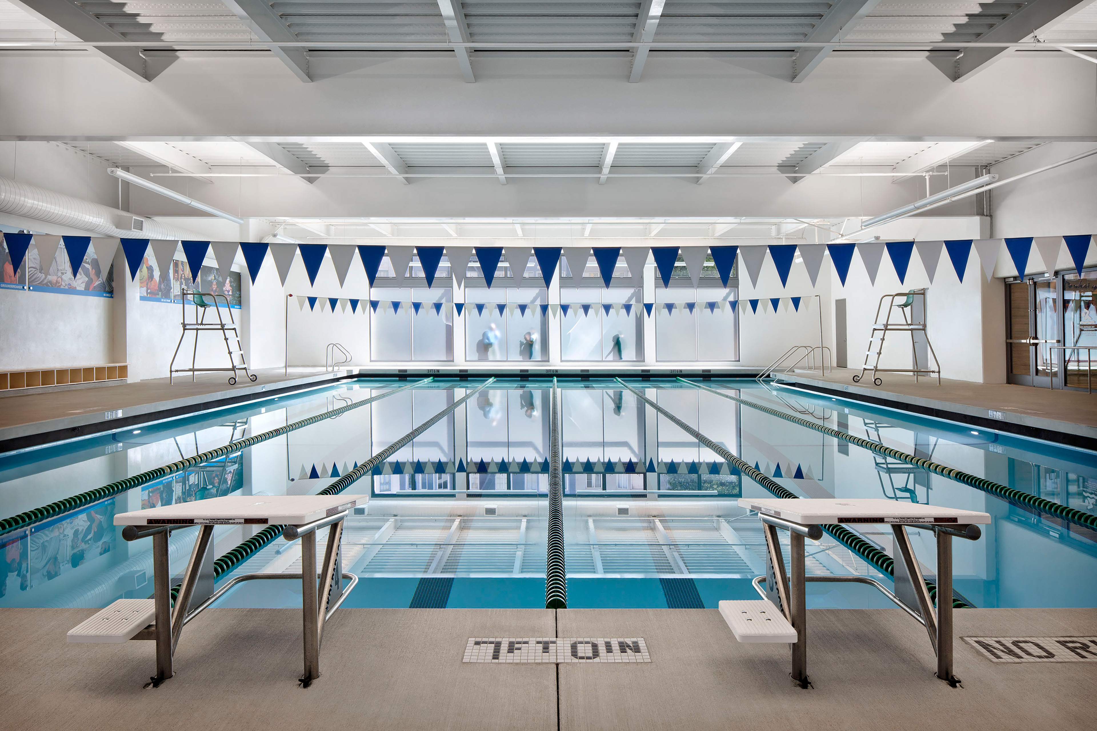 Indoor swimming pool with clear lanes, blue and white flags, and bright natural light filtering through frosted windows. The calm, reflective water emphasizes the clean, modern facility design.