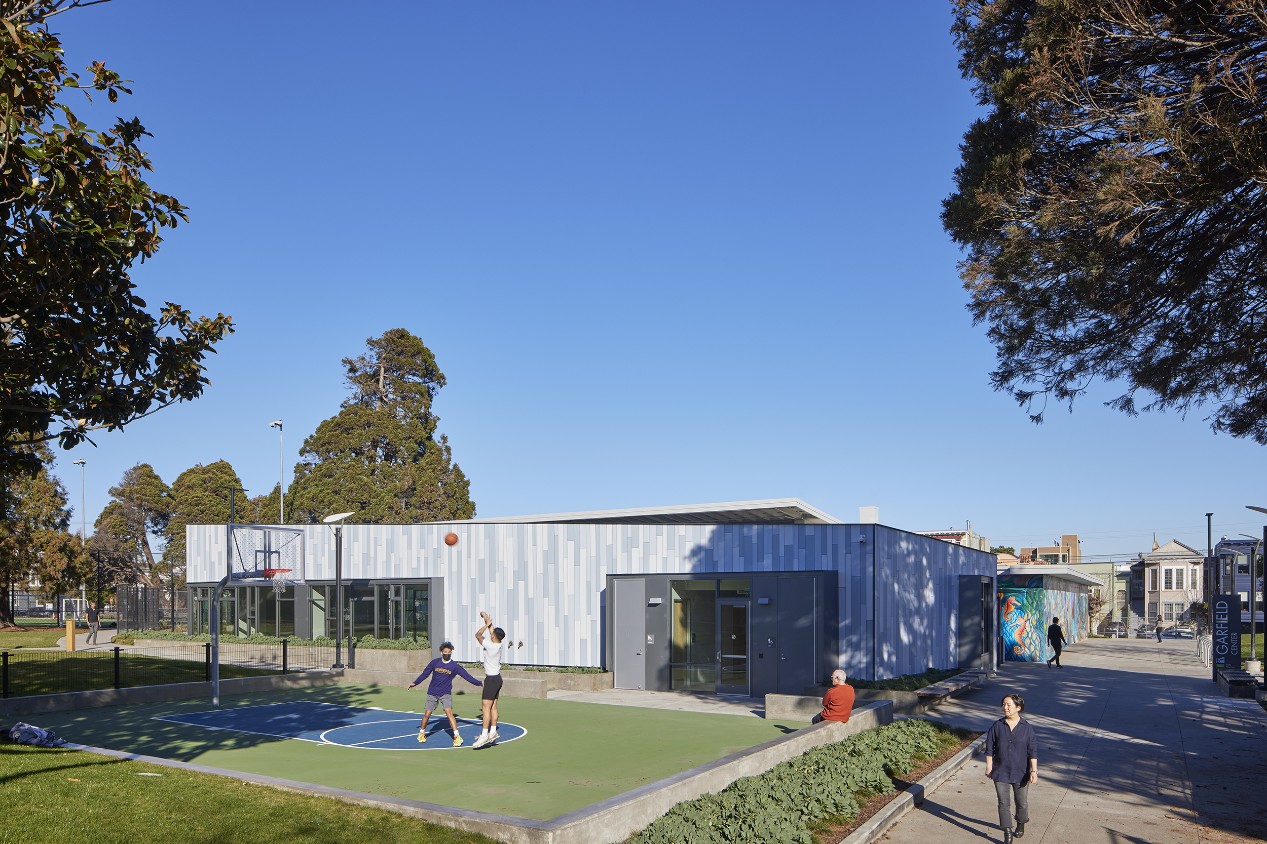 Exterior view of the Garfield Center in San Francisco, showing people enjoying the outdoor basketball court beside the modern recreation building with blue and gray paneling. Trees and nearby houses are visible under a clear blue sky.