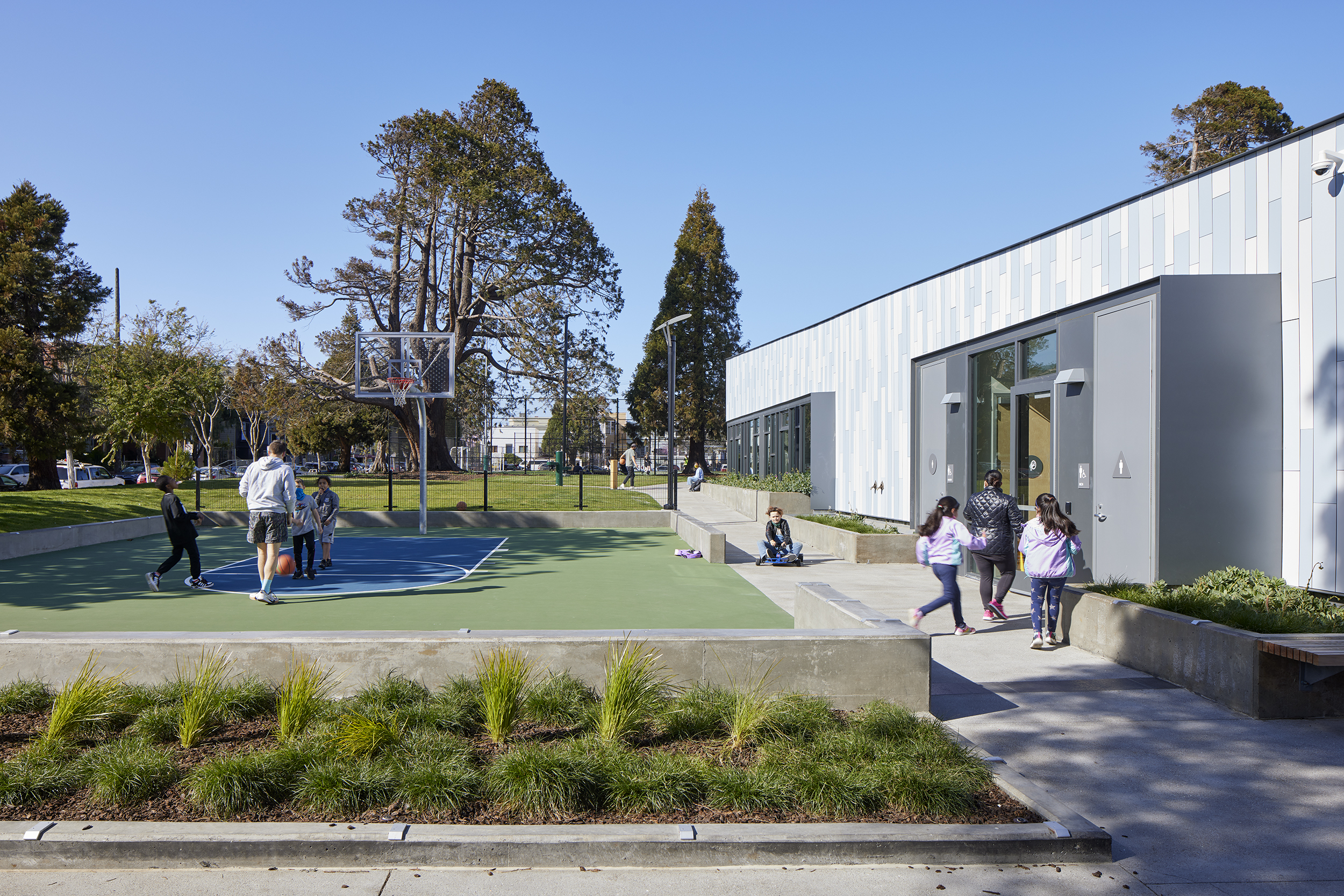 Exterior courtyard featuring a small basketball court where children play under a clear blue sky. To the right a modern clubhouse building has blue tone cladding, large windows, and landscaped planters with greenery. Restroom entrances are visible through the windows.