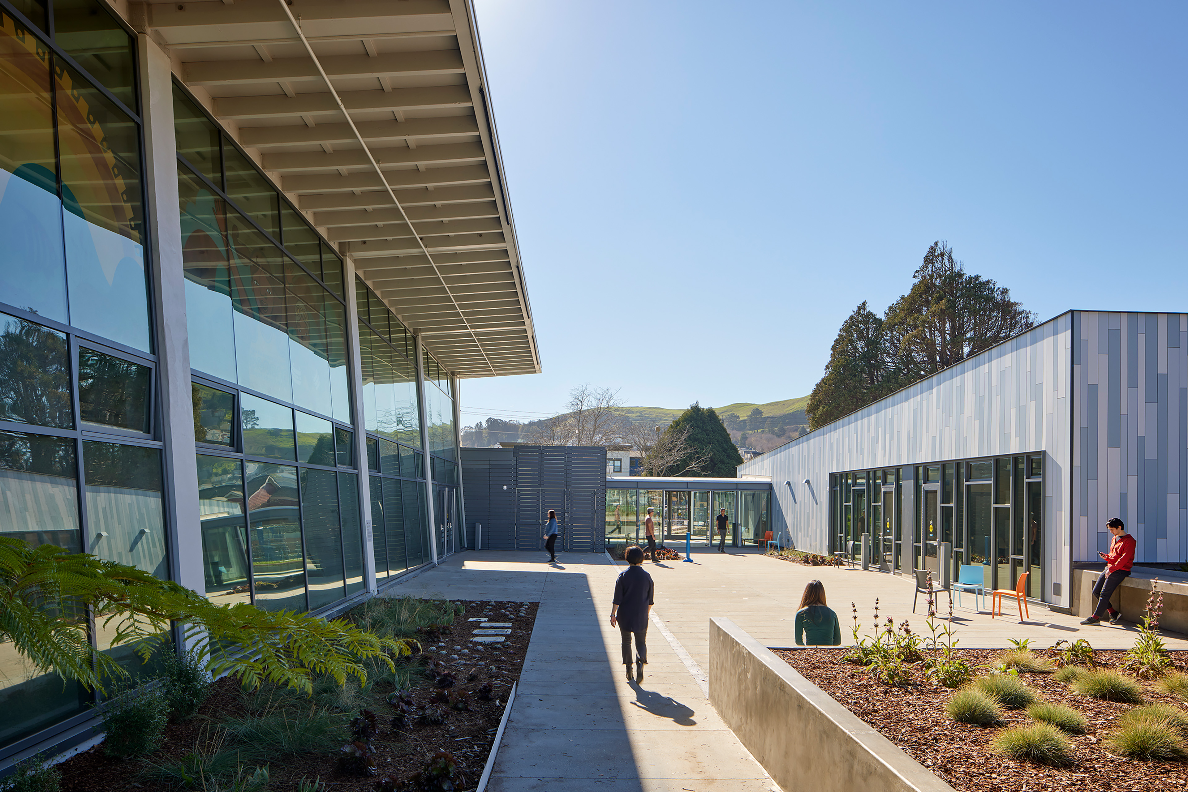 Exterior courtyard between two recreation buildings, with glass walls, metal cladding, landscaped planting beds, and outdoor seating. People move through and pause in the sunny plaza, with green hills and trees visible beyond the site.