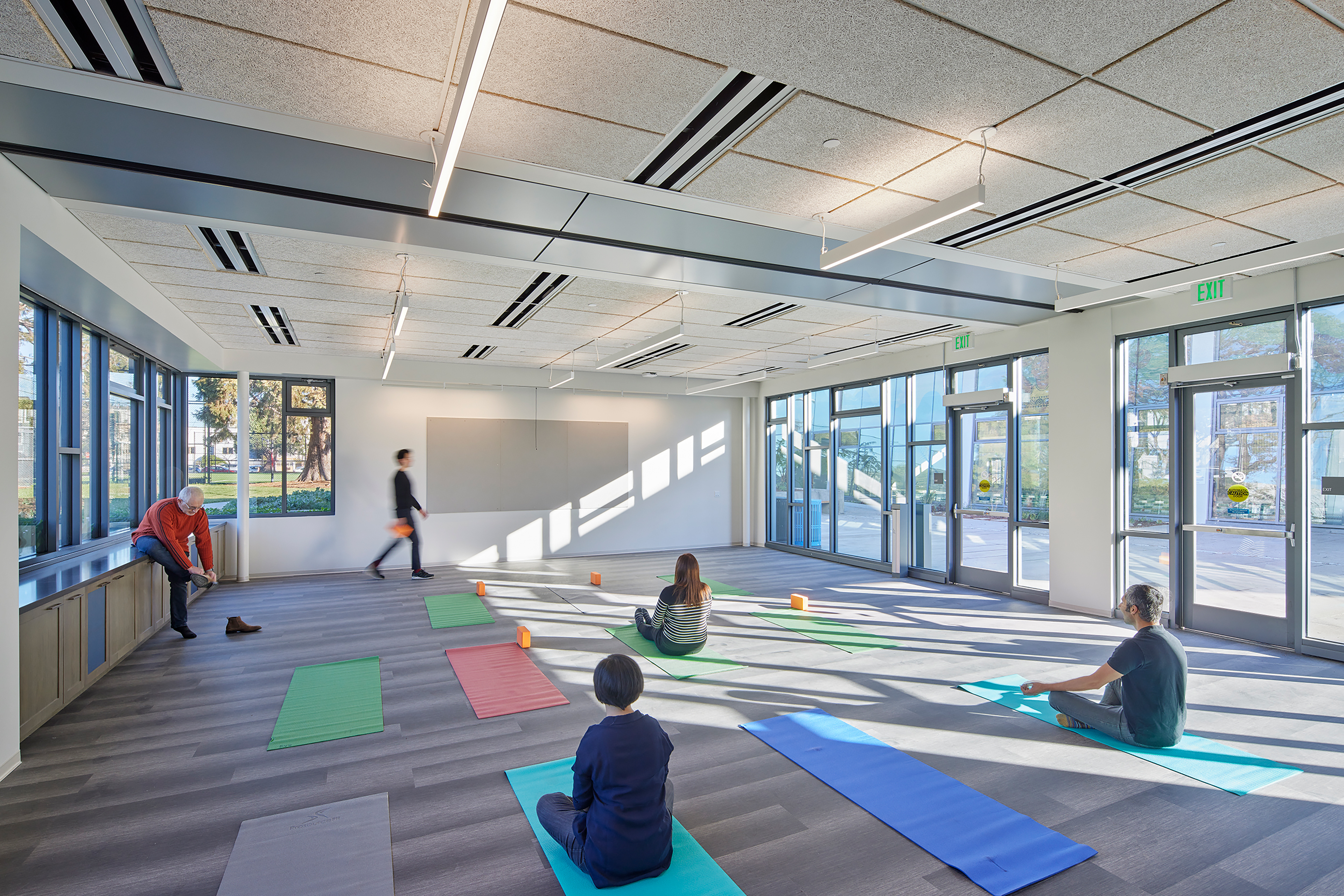 Interior of a bright, sunlit multipurpose room at the Garfield Center with people participating in a yoga or meditation class. Colorful mats are spread across the floor, and large windows let in abundant natural light.