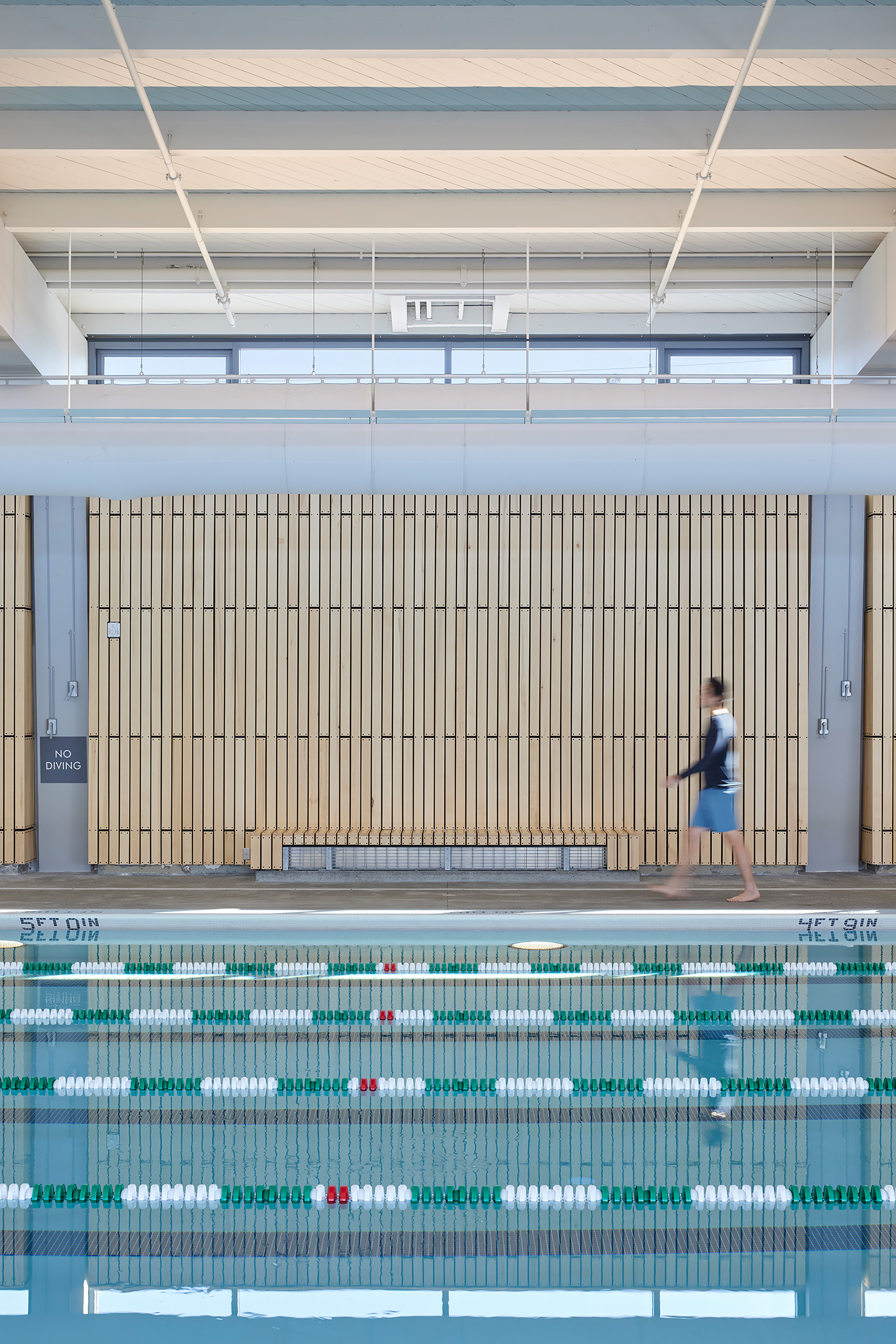 Indoor lap pool with green-and-white lane dividers, exposed structural beams and mechanical systems overhead, and a wood-slat wall along the pool deck. A person walks beside the pool, reflected in the water.