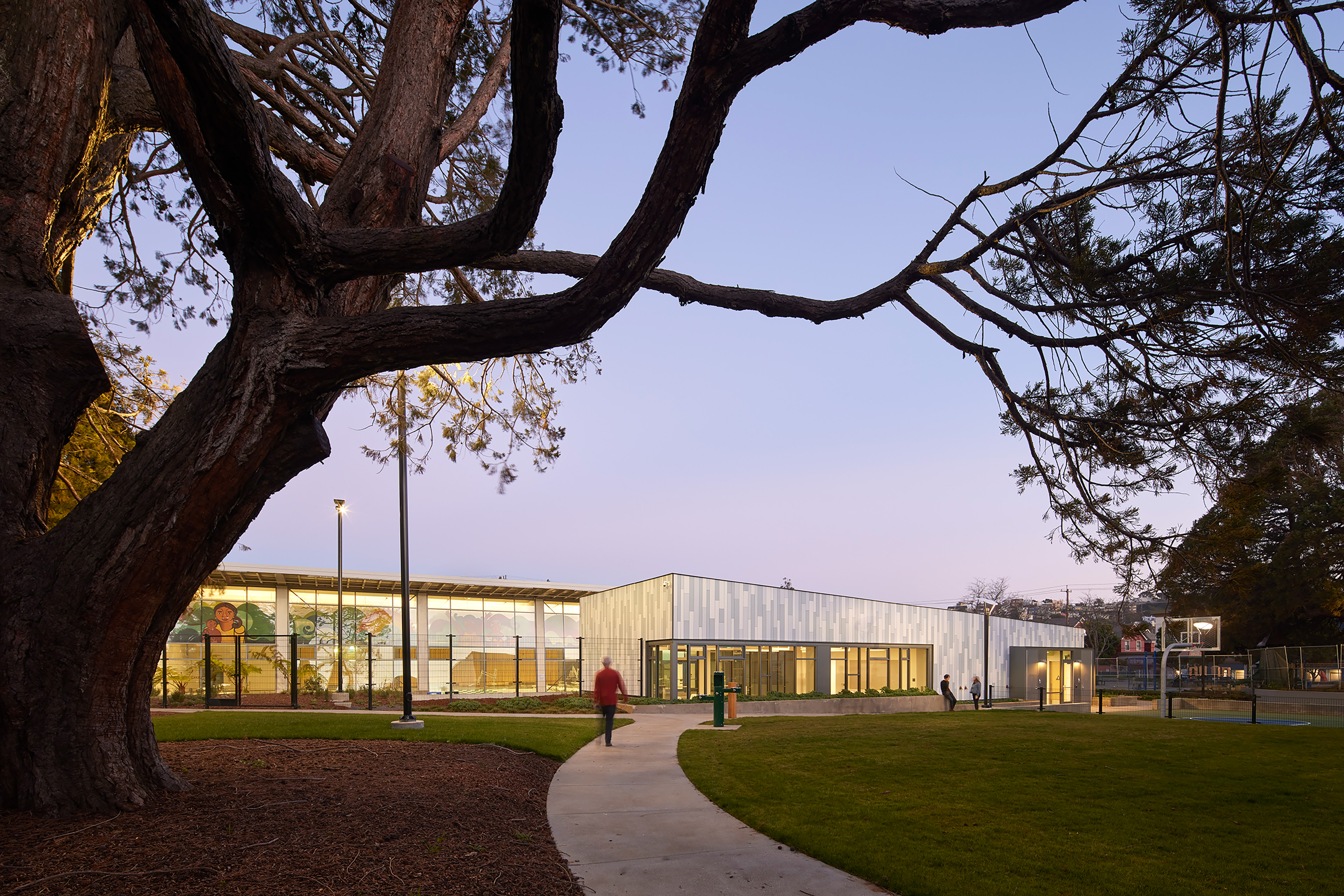 Evening view of the new recreation center with a light gray and blue exterior, seen through the branches of a large tree. Warm interior lights glow through expansive windows as people walk along a curved path across the surrounding lawn.