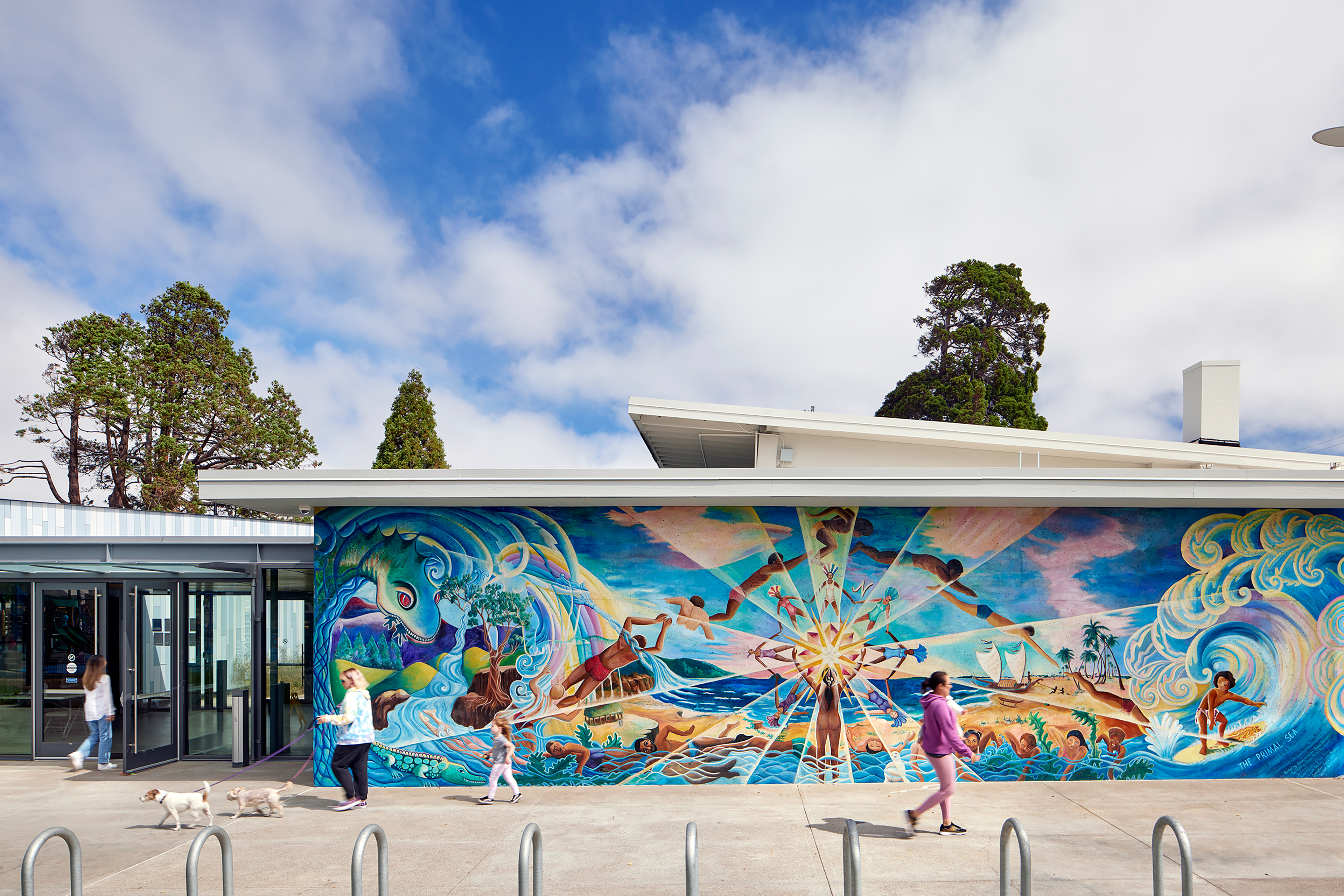 Exterior view of a modern community building with a vibrant mural depicting ocean waves, people, and nature scenes. Several people walk by, including a woman with two dogs, under a bright blue sky with scattered clouds.