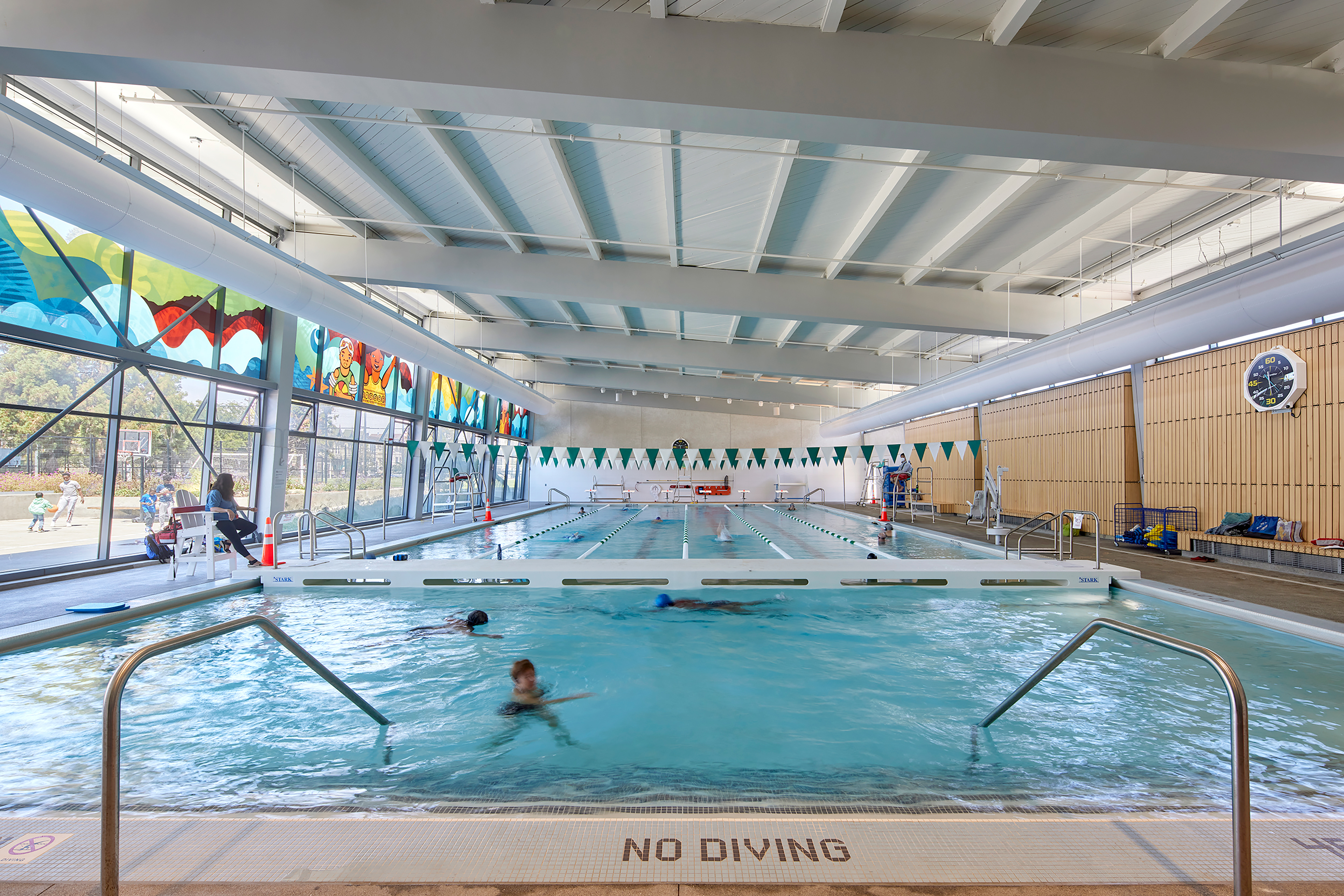 Indoor swimming pool with several swimmers in the water and a lifeguard seated along the pool’s edge. The space features large windows with colorful murals, wood slat wall panels, and bright natural light. A bulkhead divider separates lap lanes from free swim.