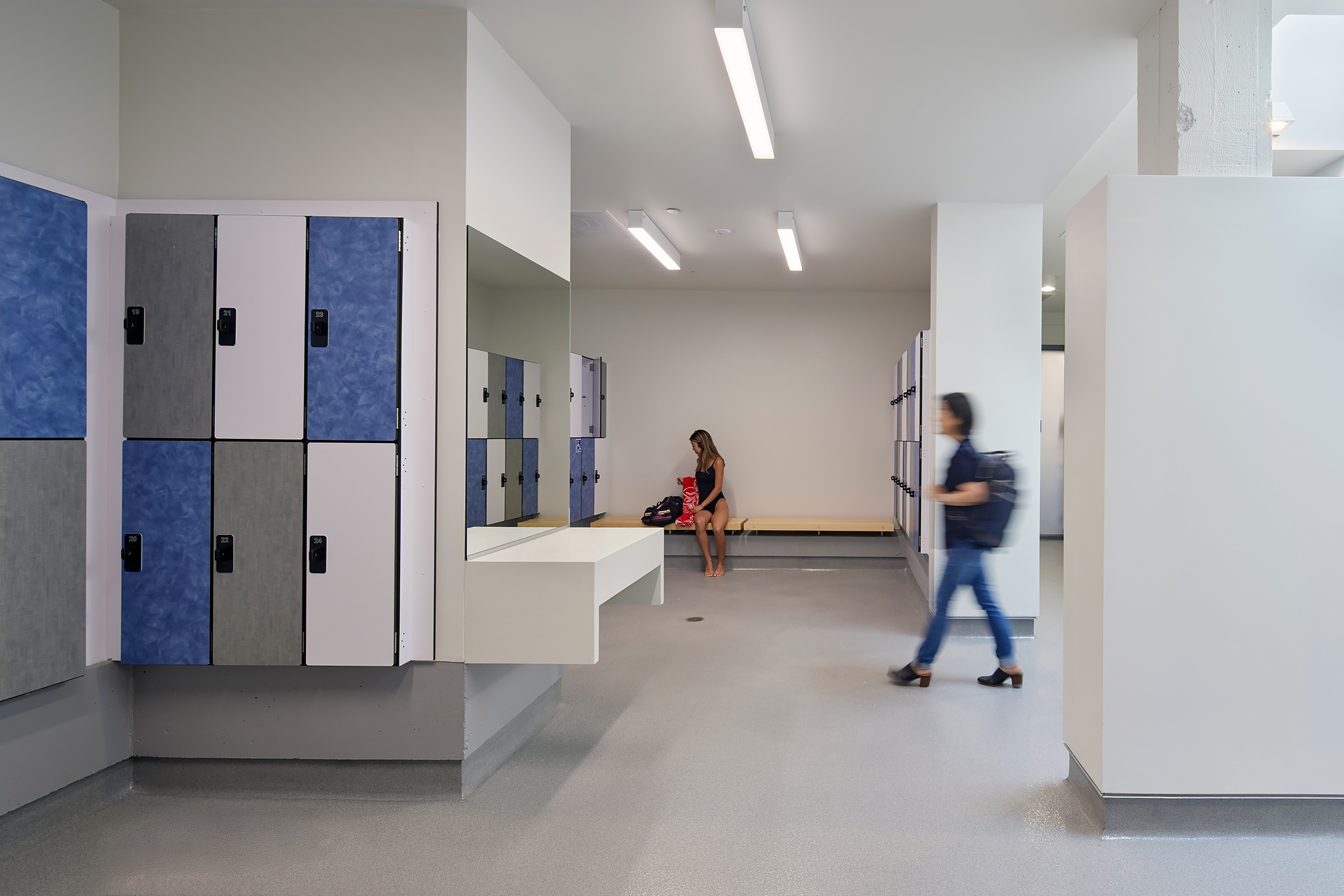 Locker room interior with blue and gray lockers, a central counter and mirror, and benches along the walls. A person in a swimsuit sits on a bench, while another walks past carrying a backpack.