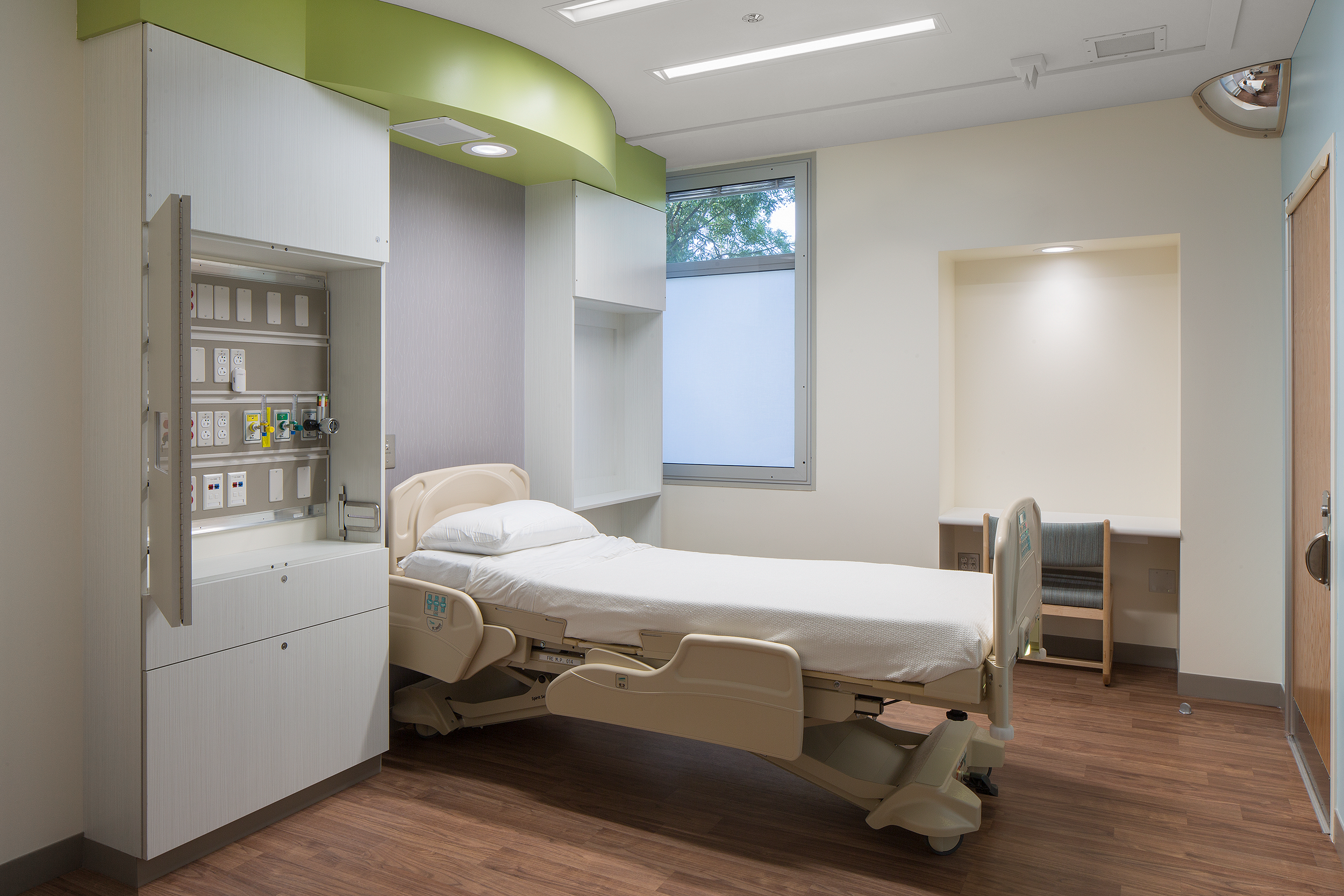Modern healthcare patient room featuring a single hospital bed, integrated medical equipment cabinet, wood-style flooring, and soft natural light filtering through a window.