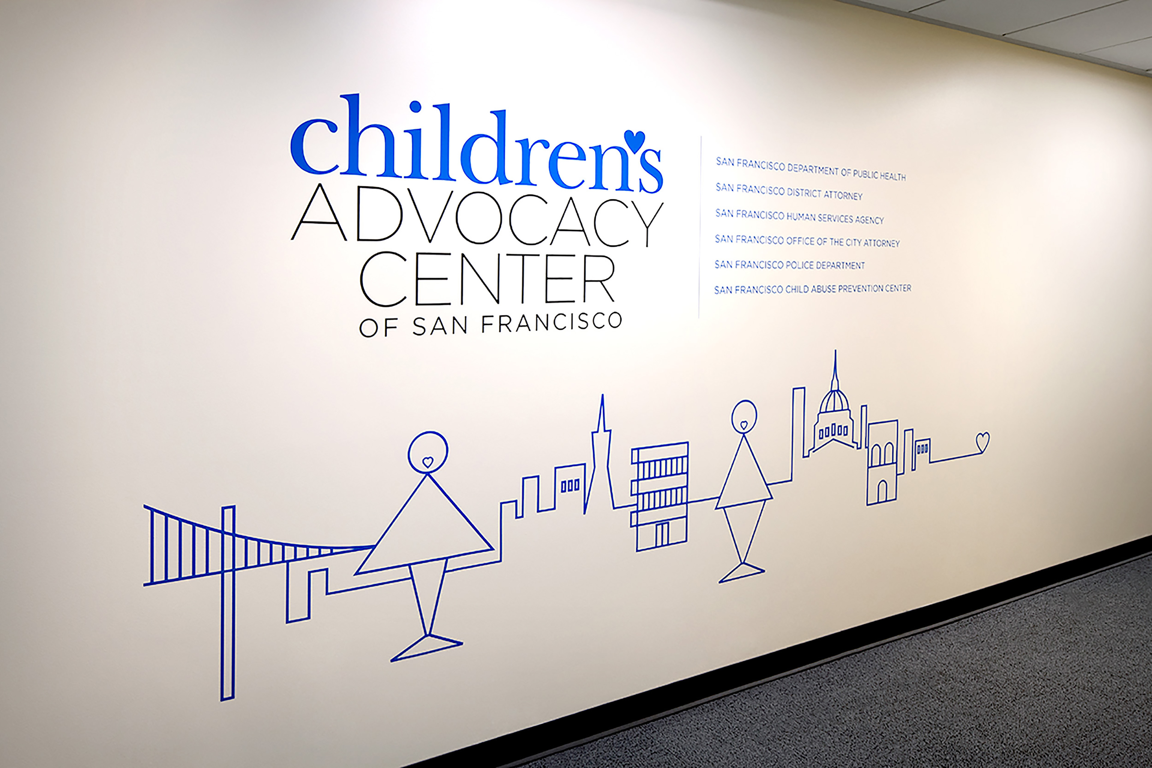 Interior wall featuring the Children’s Advocacy Center of San Francisco logo with partner organizations listed beside it. A simple blue line drawing along the wall depicts San Francisco landmarks and stylized human figures, symbolizing community connection and care.