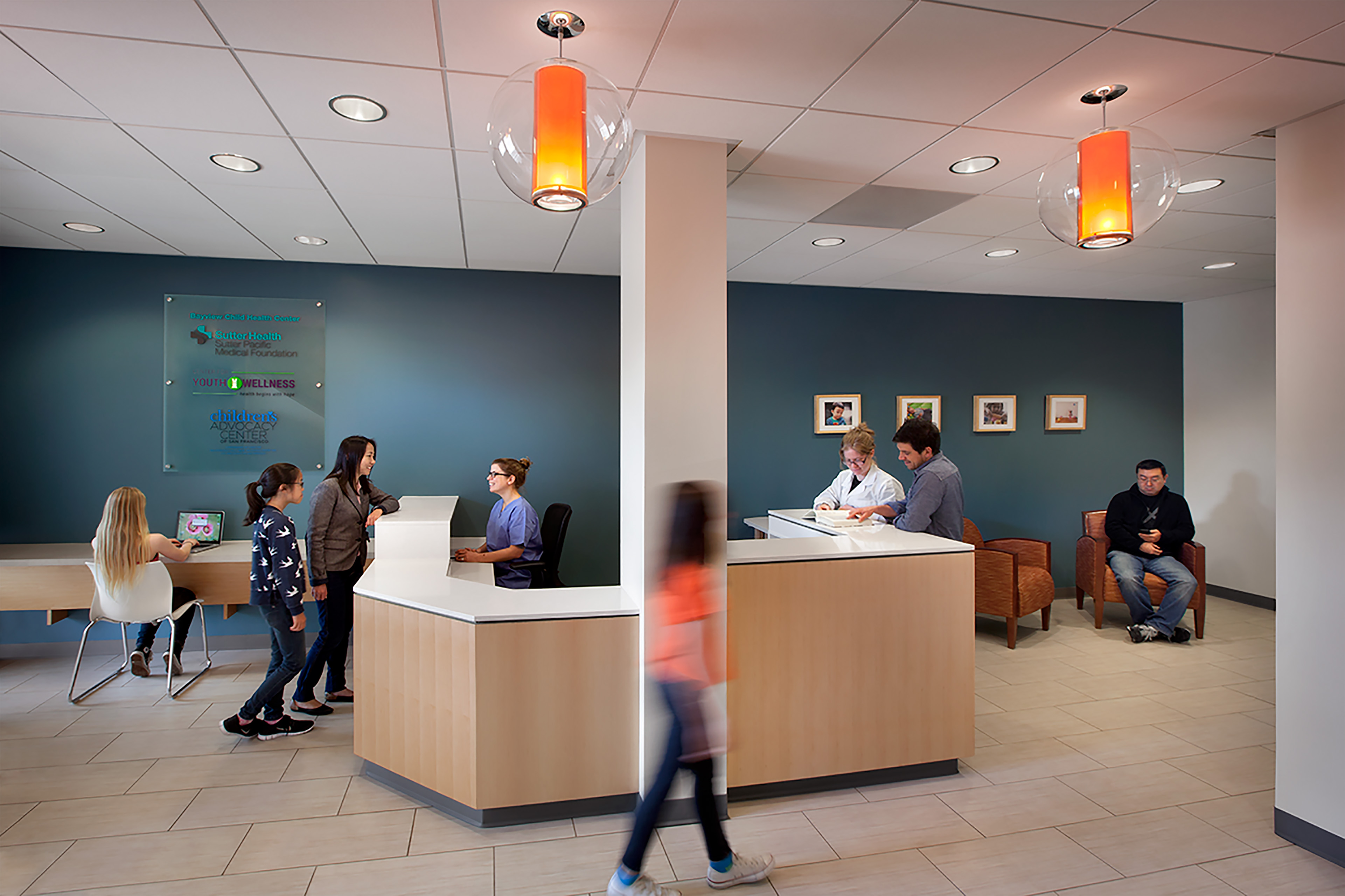 Bright and welcoming healthcare reception area with a central check-in desk and warm wood finishes. People are interacting with staff at the counter, while others sit in the waiting area or use a tablet at a side desk. The space features teal accent walls, framed artwork, modern orange pendant lights, and comfortable seating, creating a calm and inviting environment.