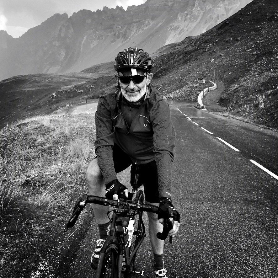 Alyosha wearing cycling gear and sunglasses riding a bicycle on a winding mountain road, captured in black and white with misty peaks in the background.