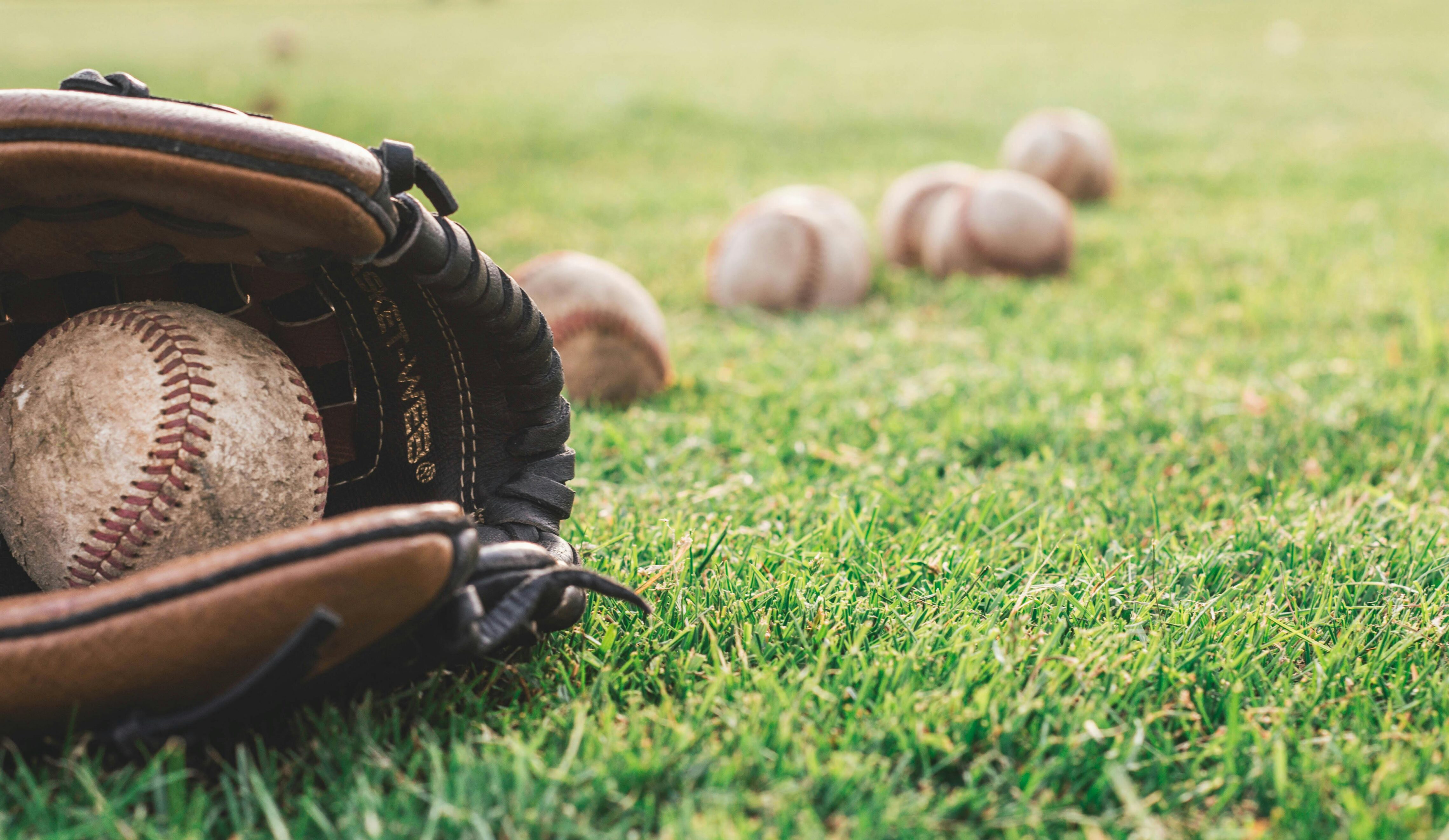 A close-up of a worn, dirty baseball resting inside a brown leather glove on green grass, with several other baseballs scattered out of focus in the background.