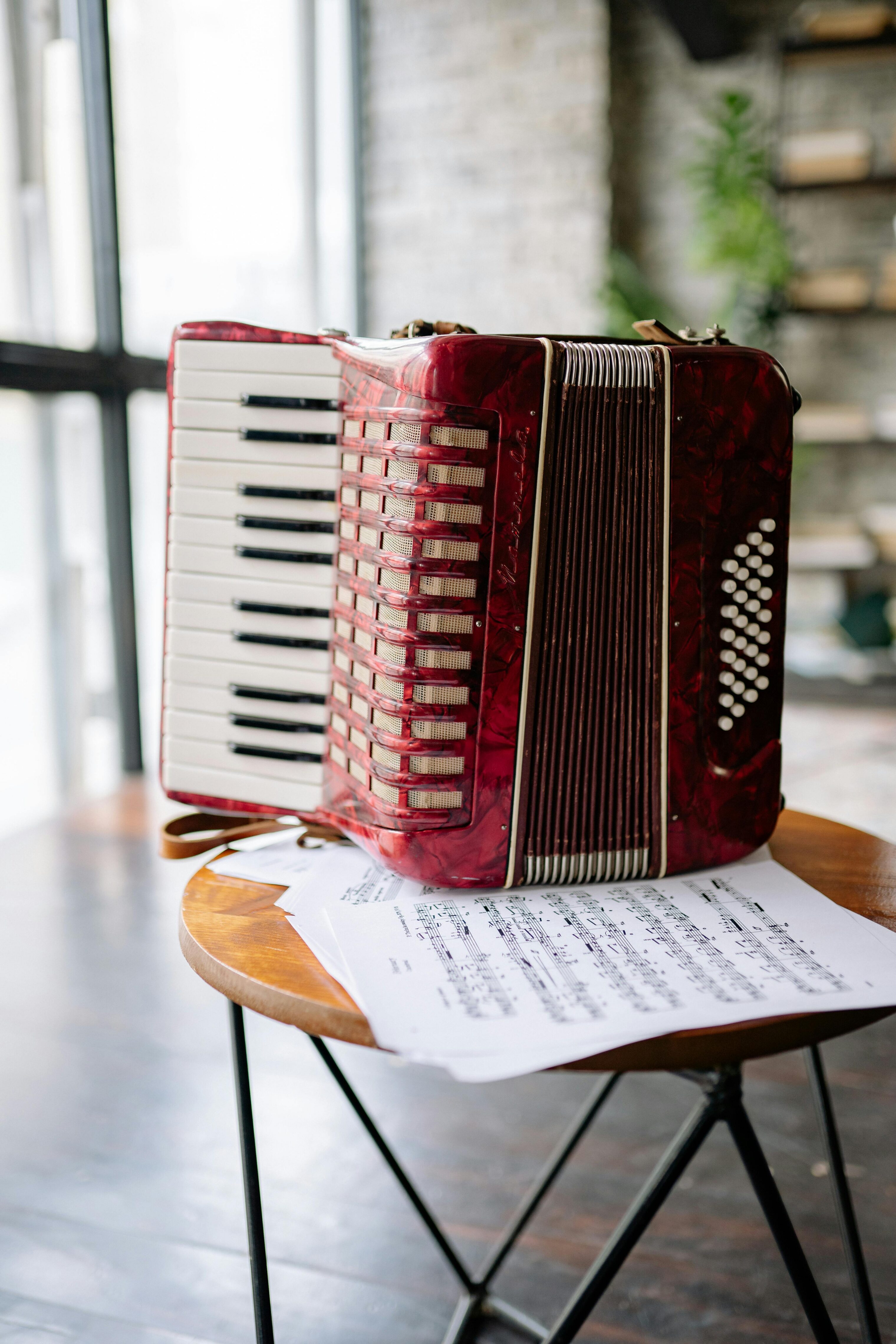Red accordion resting on a wooden table with sheet music underneath in a bright room.