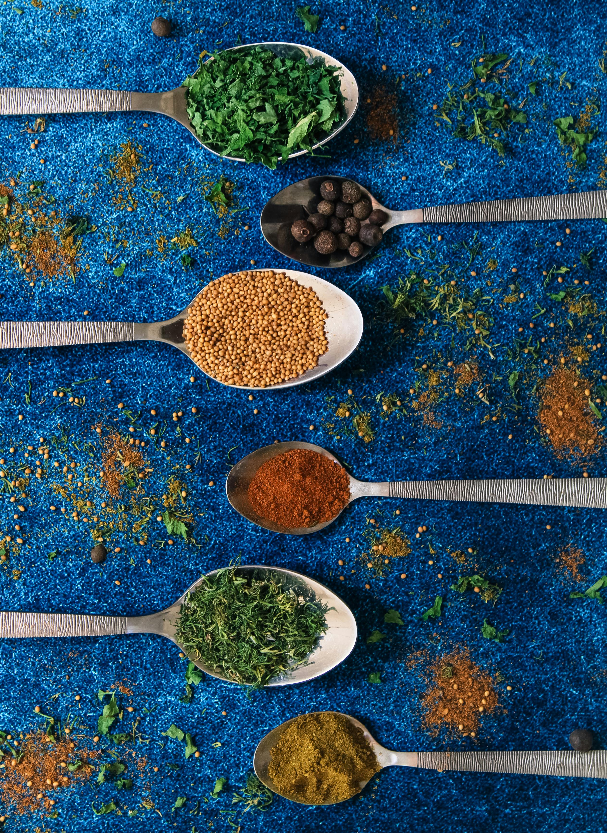 Overhead view of metal spoons filled with various herbs and spices on a blue textured surface.