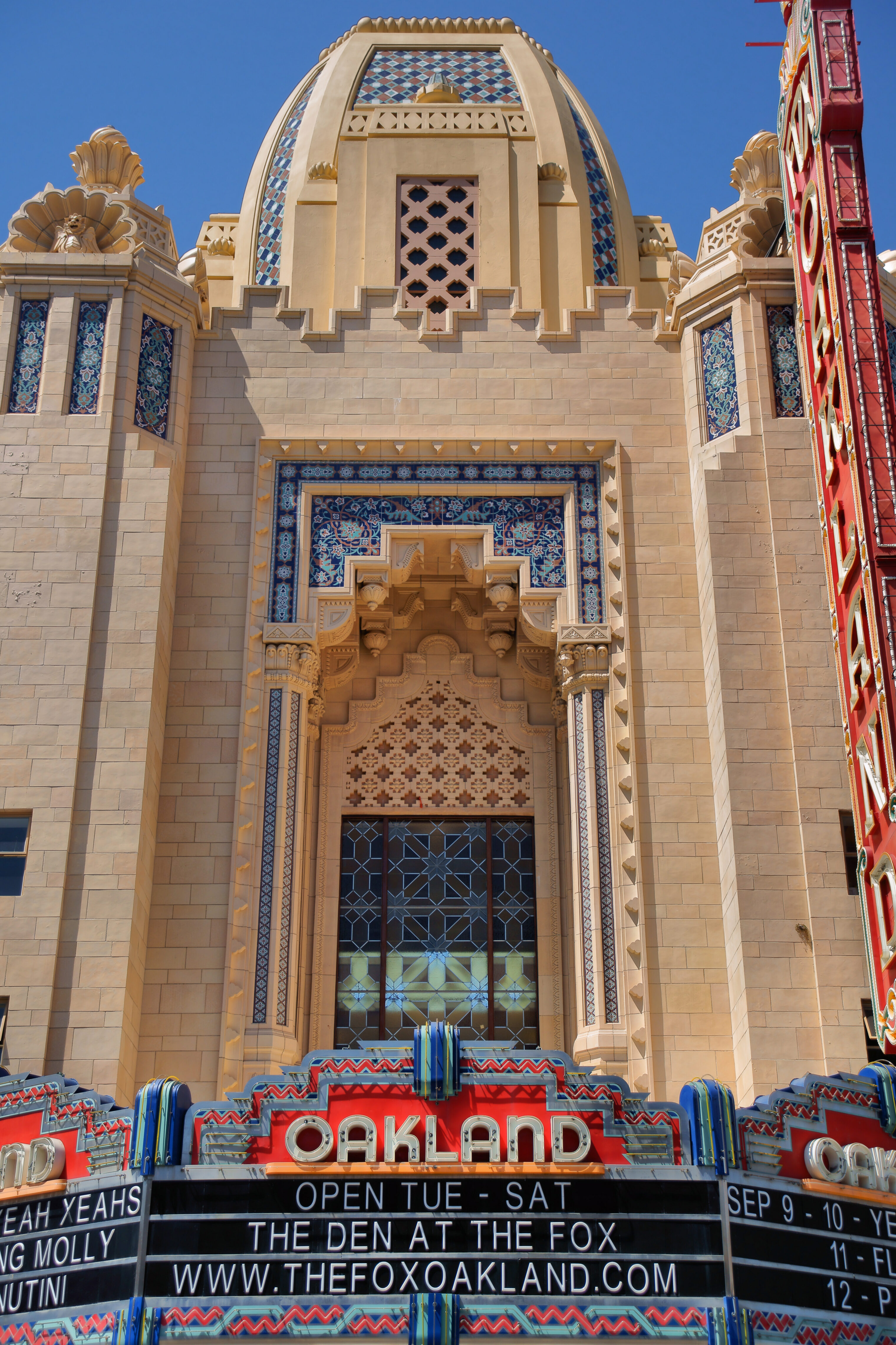 Exterior view of the Fox Theater in Oakland, California, featuring its ornate Art Deco façade with geometric tile patterns, arched window, and marquee sign reading “OAKLAND” beneath a clear blue sky.