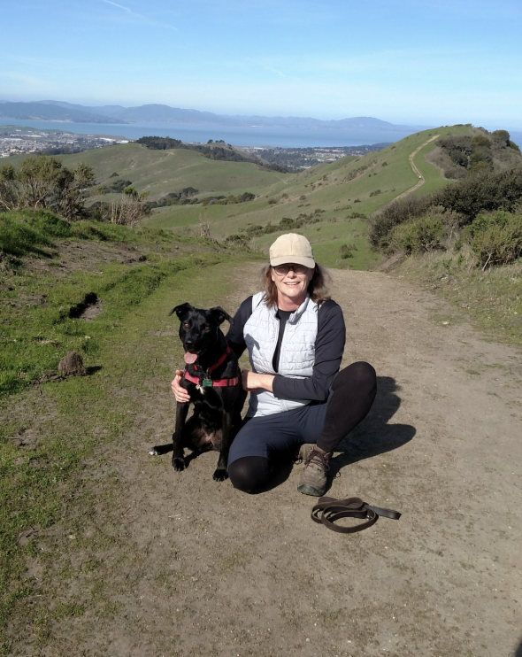 A woman kneels beside a black dog on a sunlit hillside trail, with green rolling hills, the bay, and distant mountains in the background.