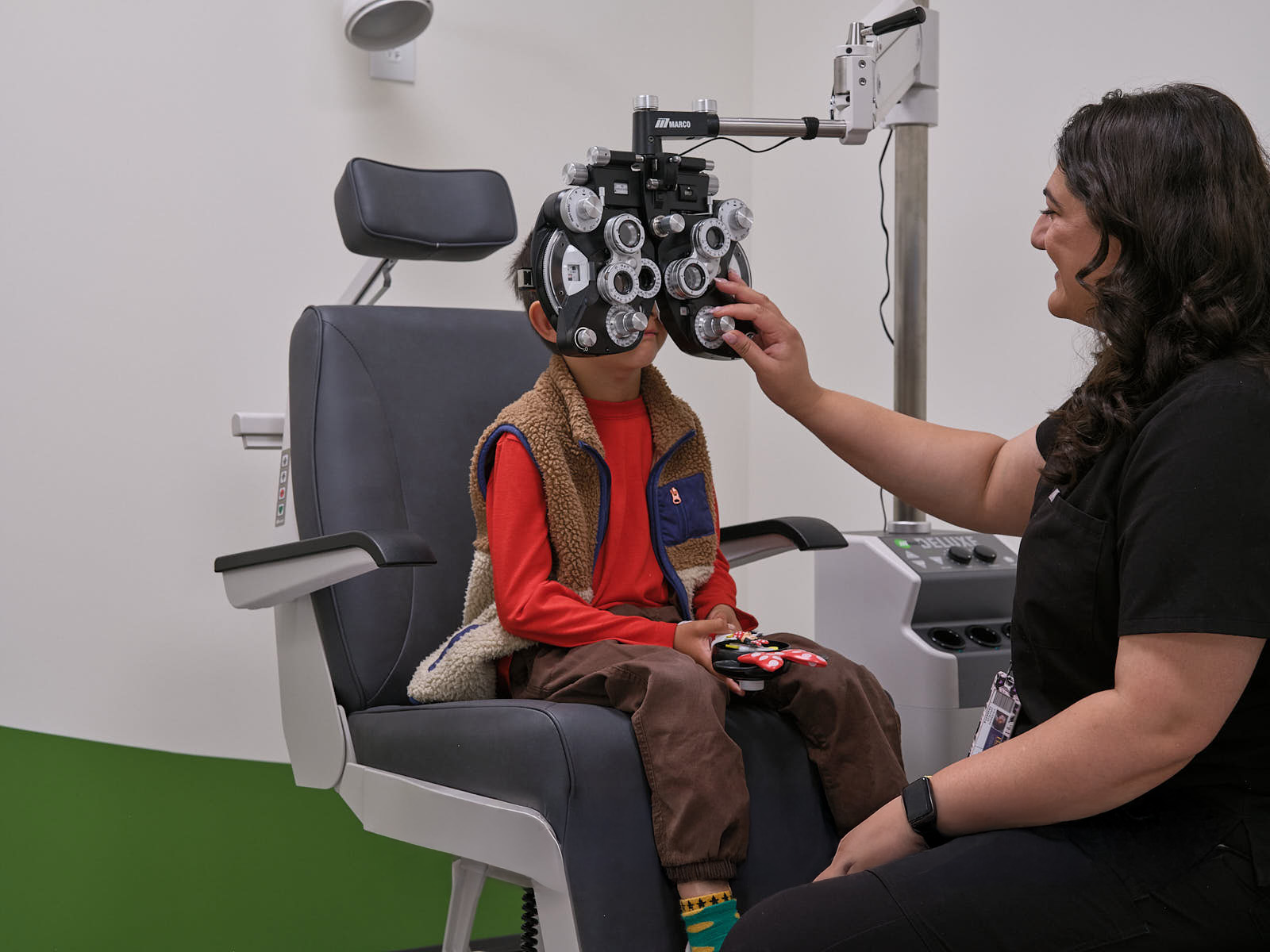 A clinician adjusts a phoropter while a child seated in an exam chair undergoes an eye examination.