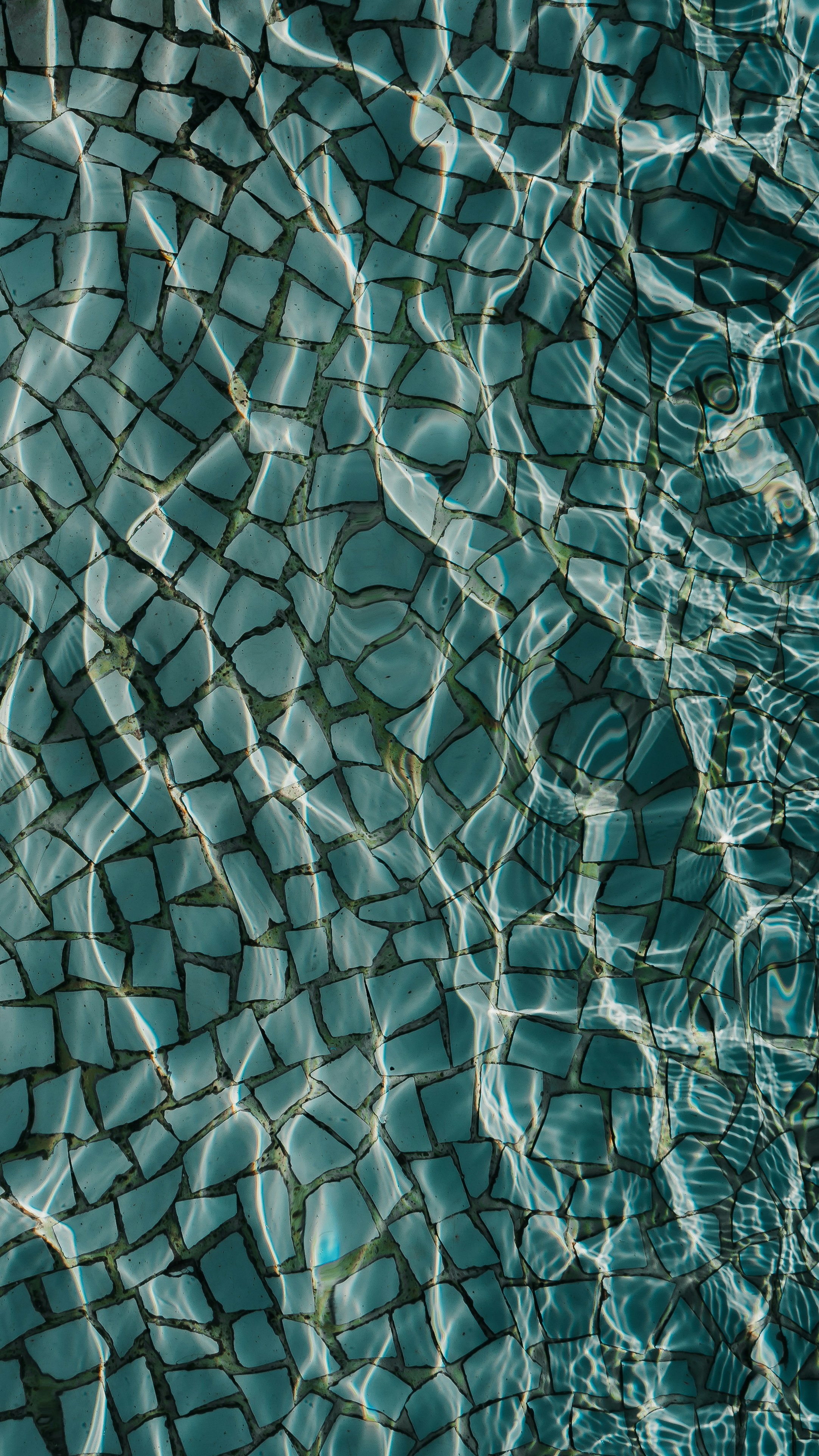 Close-up of turquoise mosaic tiles beneath rippling water, with sunlight creating shimmering reflections and patterns across the surface.