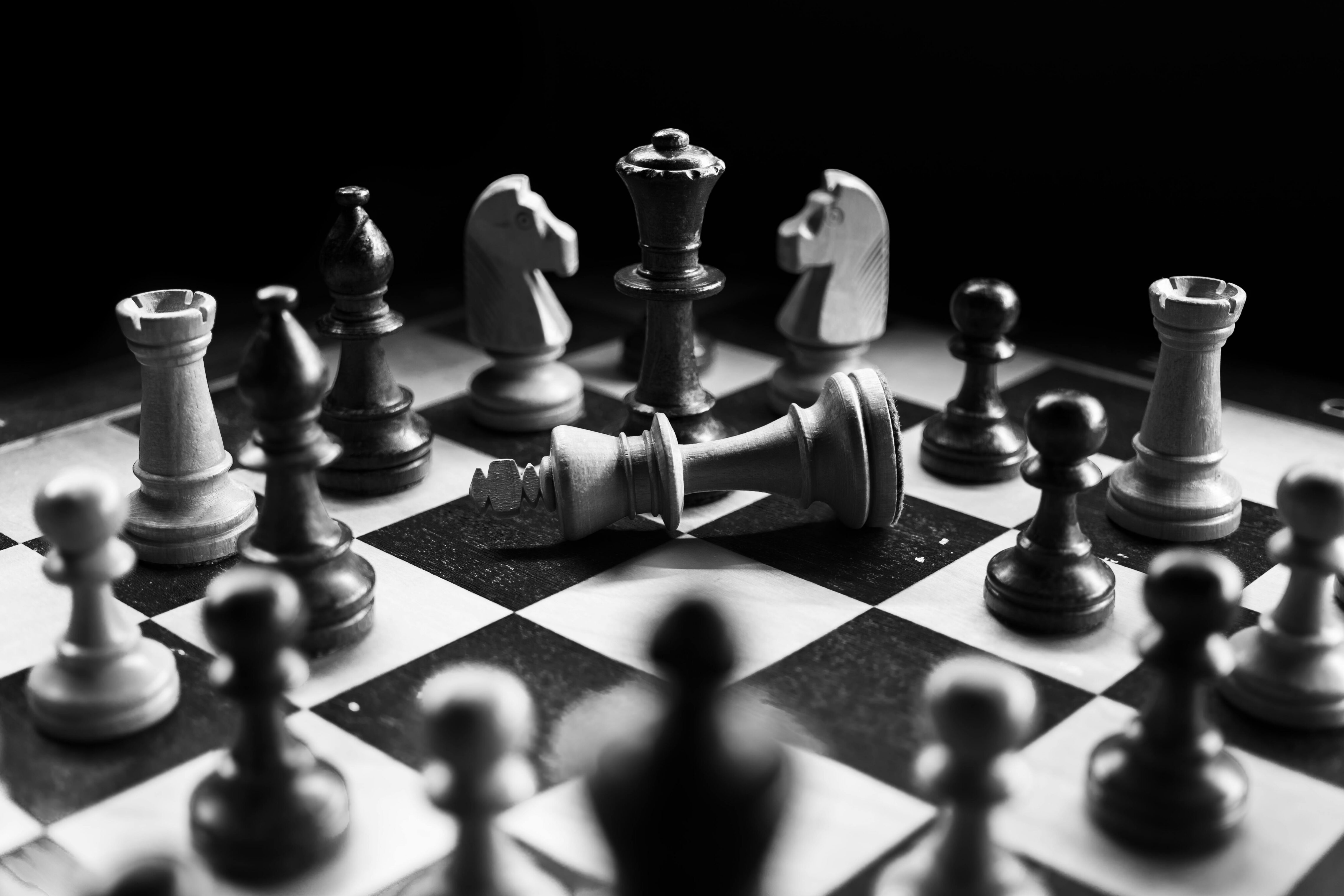 Black and white photo of a chessboard showing a dramatic checkmate moment with the white king toppled over among standing chess pieces, surrounded by both light and dark pieces under strong lighting and deep shadows.