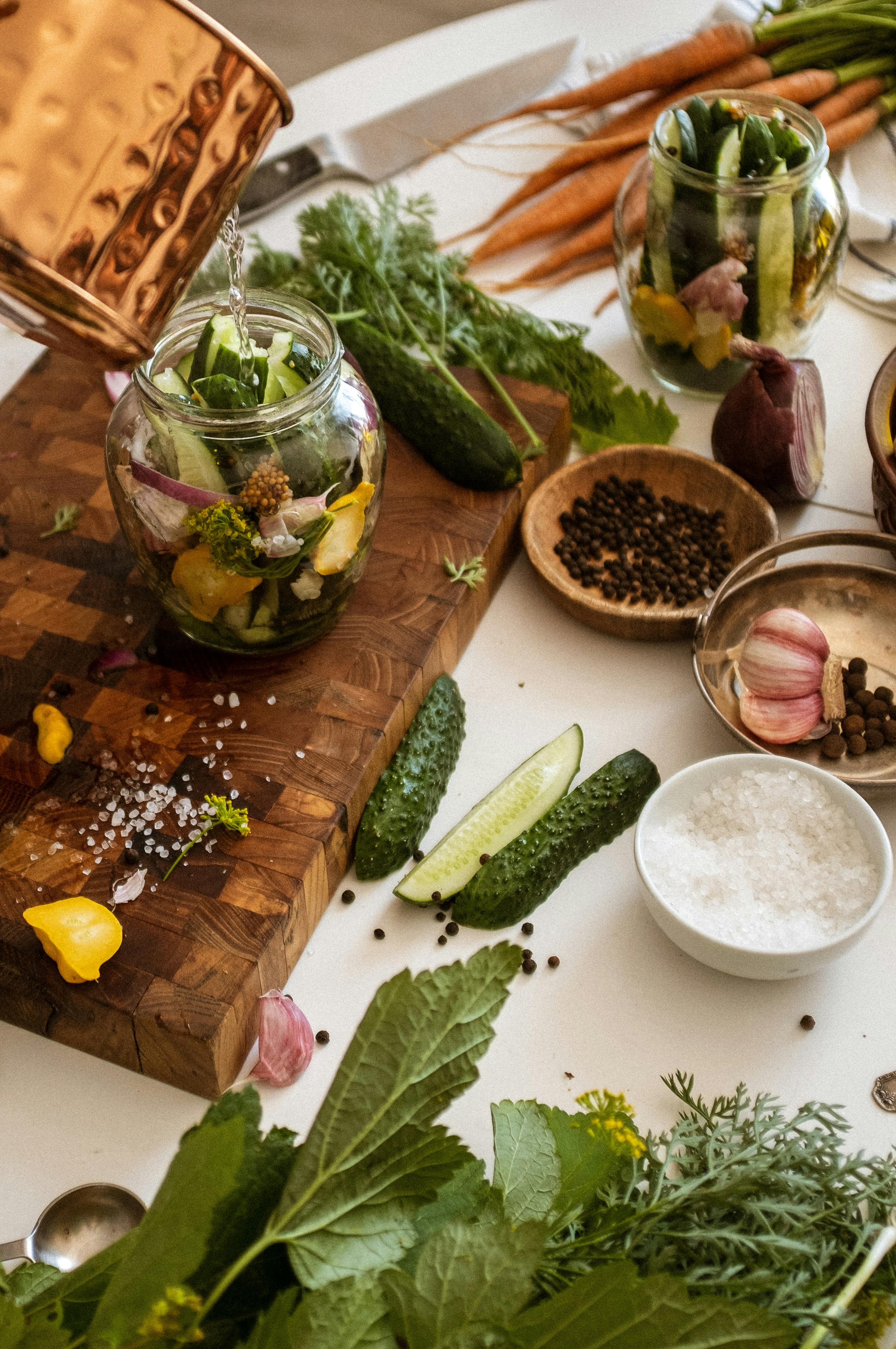 Close-up of a person preparing homemade pickles, spooning mustard seeds into a glass jar filled with sliced cucumbers, garlic, and herbs on a wooden cutting board.