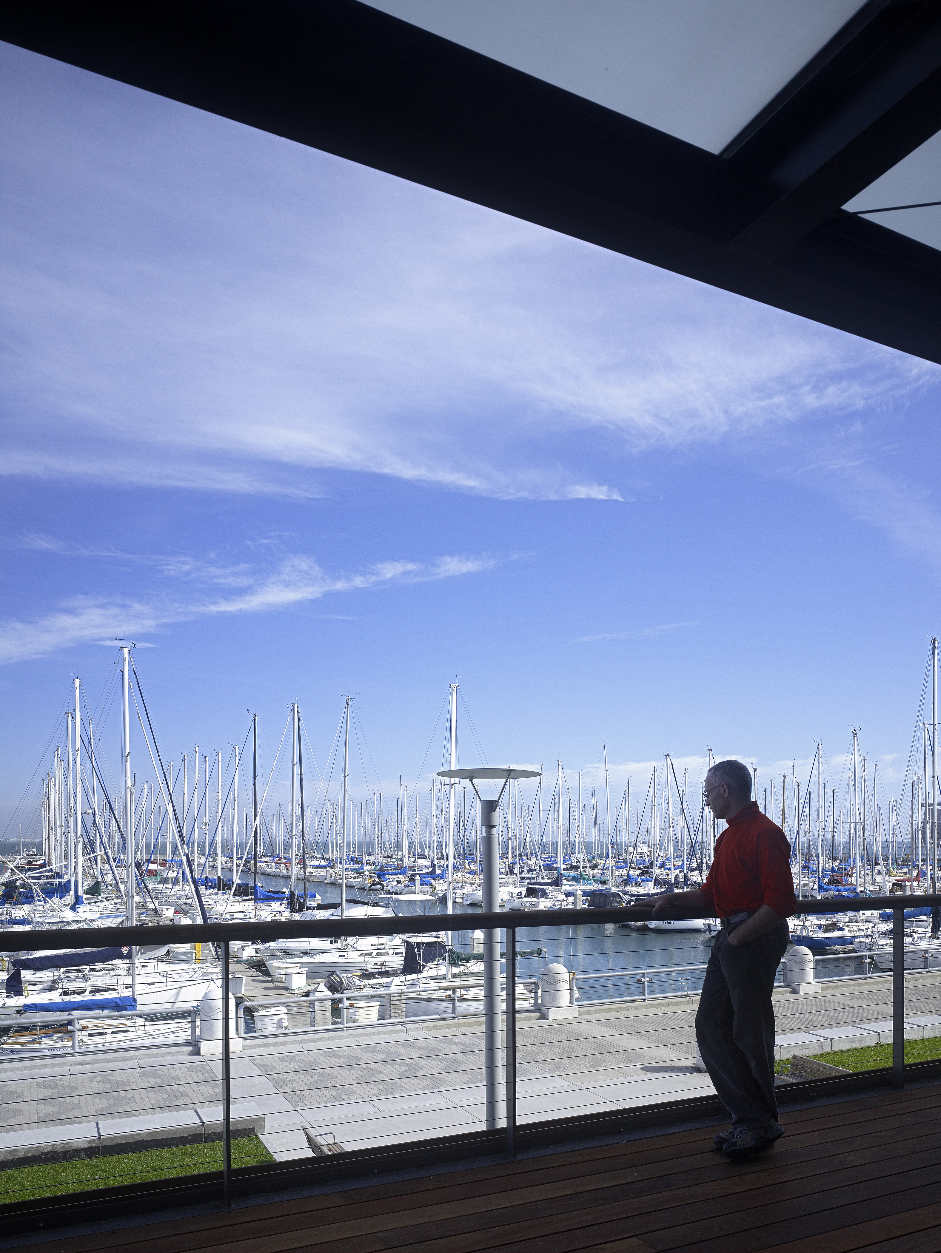 Person standing on an upper-level terrace, leaning against a railing and looking out over a marina filled with sailboats, with blue sky and wispy clouds beyond.