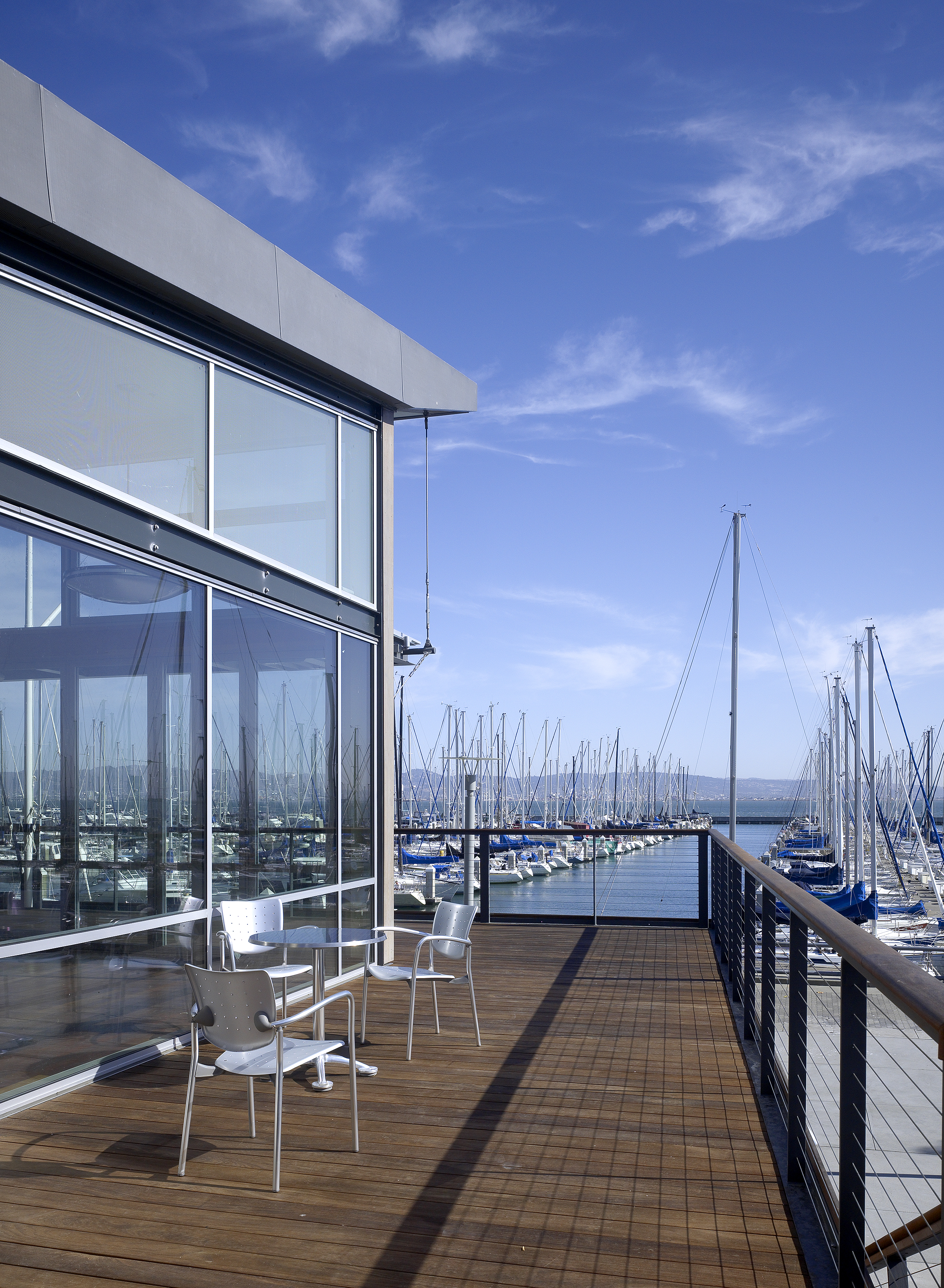 A wood-decked terrace overlooks a marina filled with sailboats, with metal chairs and a small table positioned beside a glass-walled building under a clear blue sky.