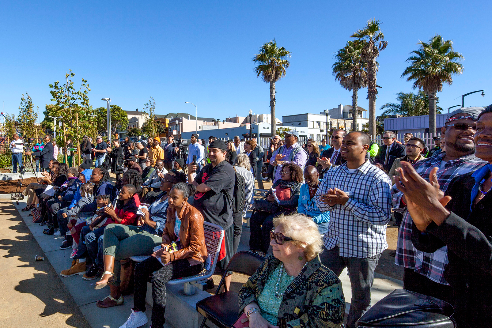 Outdoor amphitheater filled with community members seated and standing under a clear blue sky. The crowd applauds during a public event, with palm trees, plantings, and neighborhood buildings in the background.
