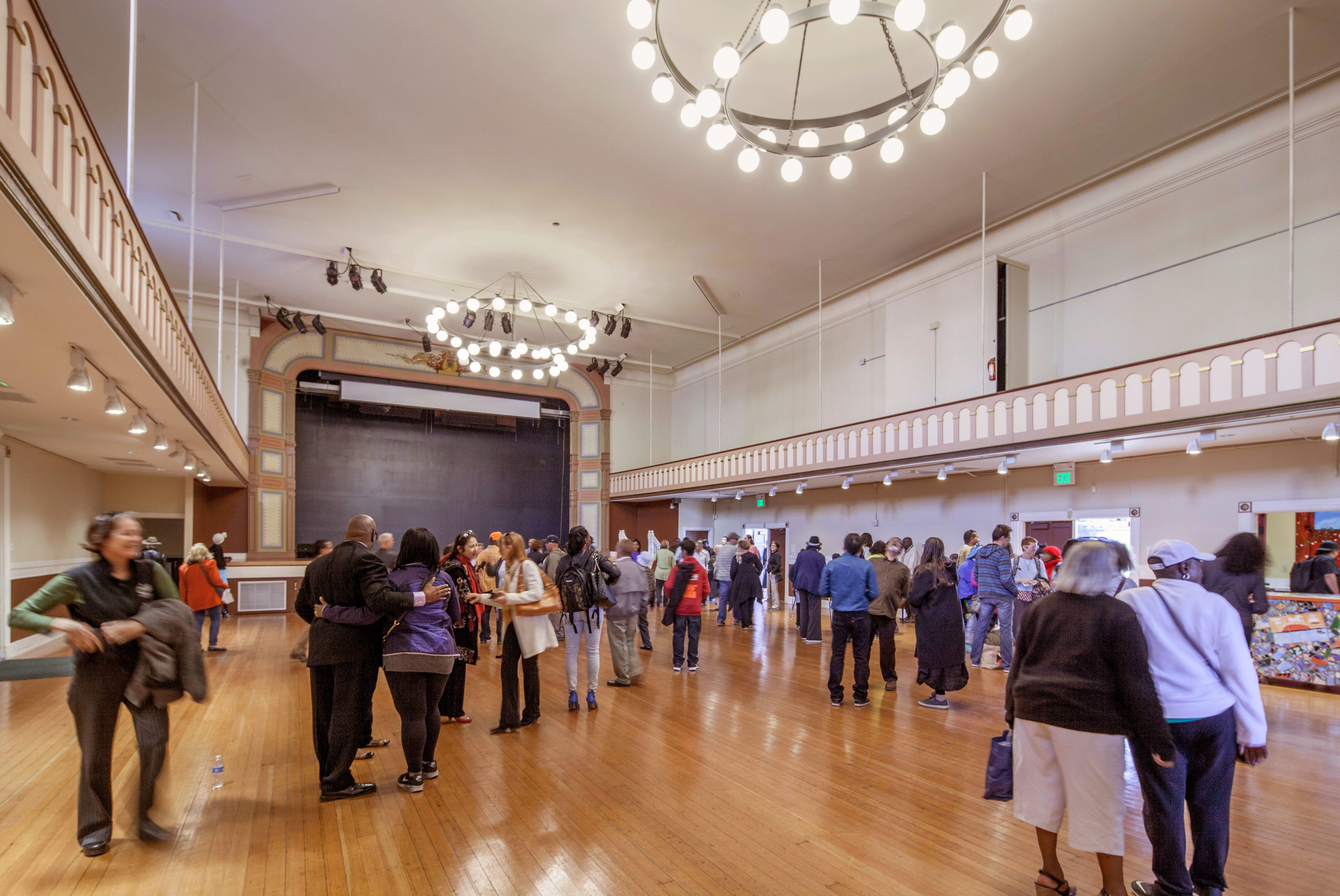 Historic auditorium interior with polished wood floors, decorative balcony railings, ring-shaped chandeliers, and a stage framed by ornate architectural detailing. Visitors gather and mingle throughout the restored event hall.