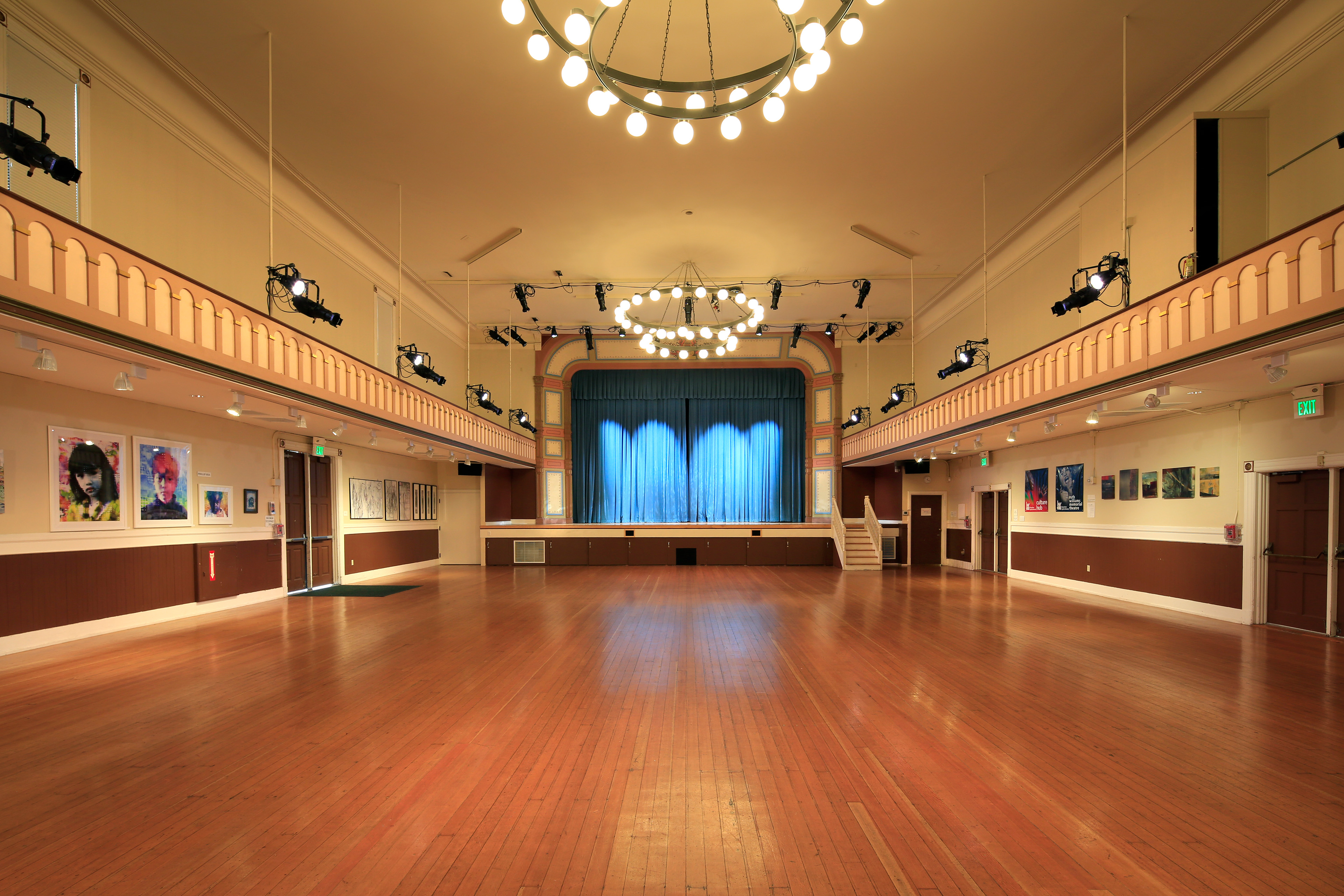 Historic auditorium with polished wood floors, a raised stage with blue curtains, decorative balconies, gallery artwork, and suspended circular chandeliers and theater lighting.
