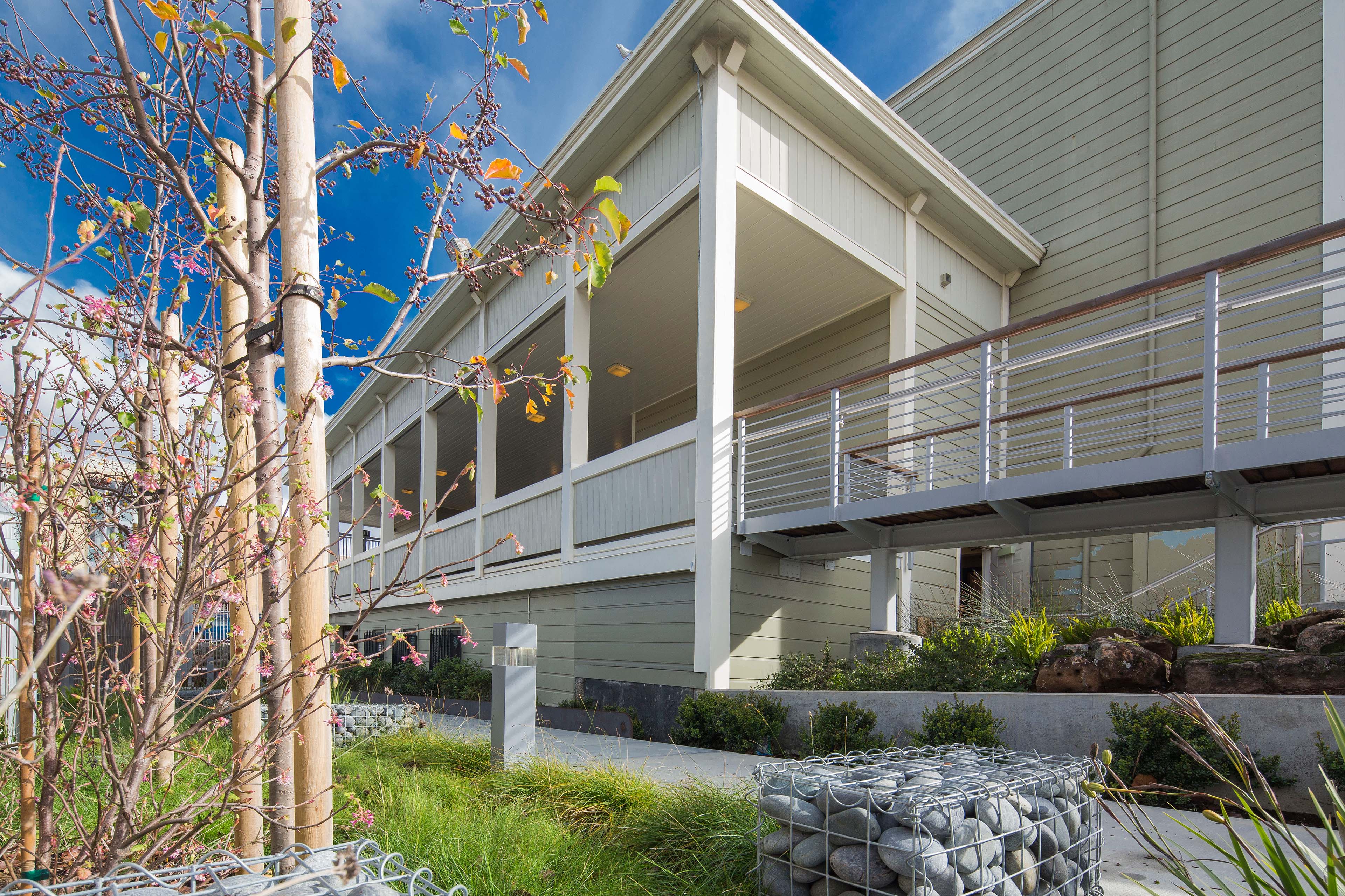 Exterior view of a school building with covered walkways, horizontal siding, metal railings, and planted landscape beds with young trees, grasses, rocks, and gabion-style stone baskets.