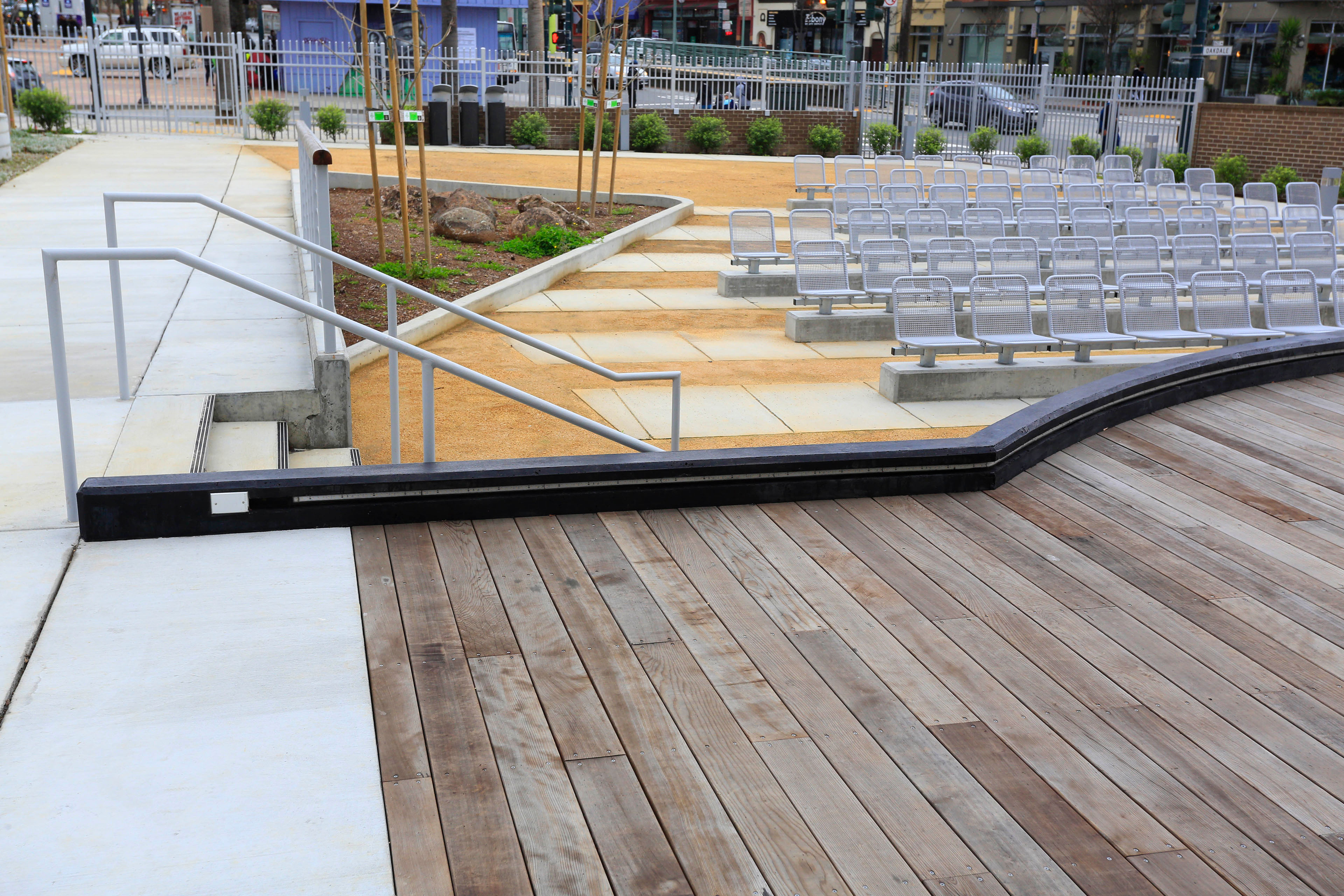 Outdoor amphitheater with a wood stage, concrete walkways, metal railings, landscaped planting areas, and rows of fixed metal seats facing the performance area.