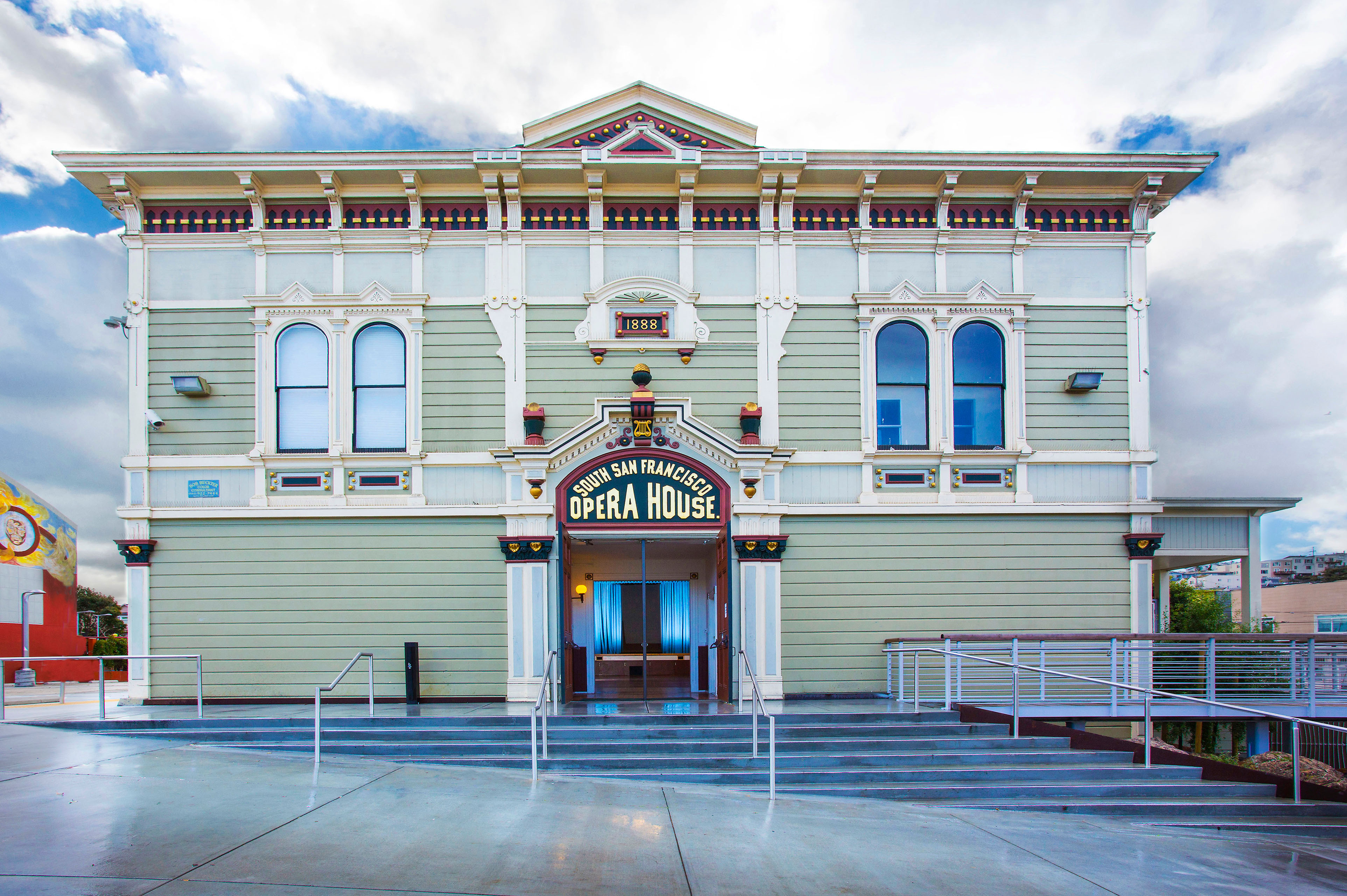 South San Francisco Opera House exterior with decorative Victorian-style detailing, arched windows, a central entrance, stairs, ramps, and metal railings. The restored façade displays an “1888” marker and “South San Francisco Opera House” sign beneath a cloudy sky.
