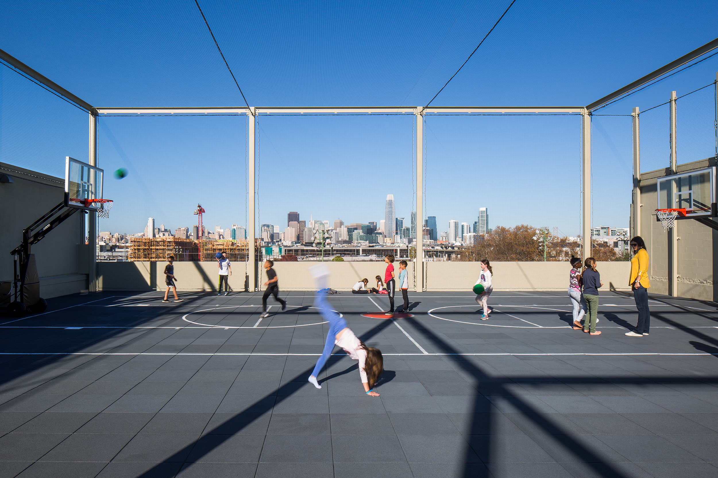 Children play and run on a rooftop outdoor basketball court enclosed by safety netting, with two hoops and a panoramic city skyline visible in the background under a clear blue sky.
