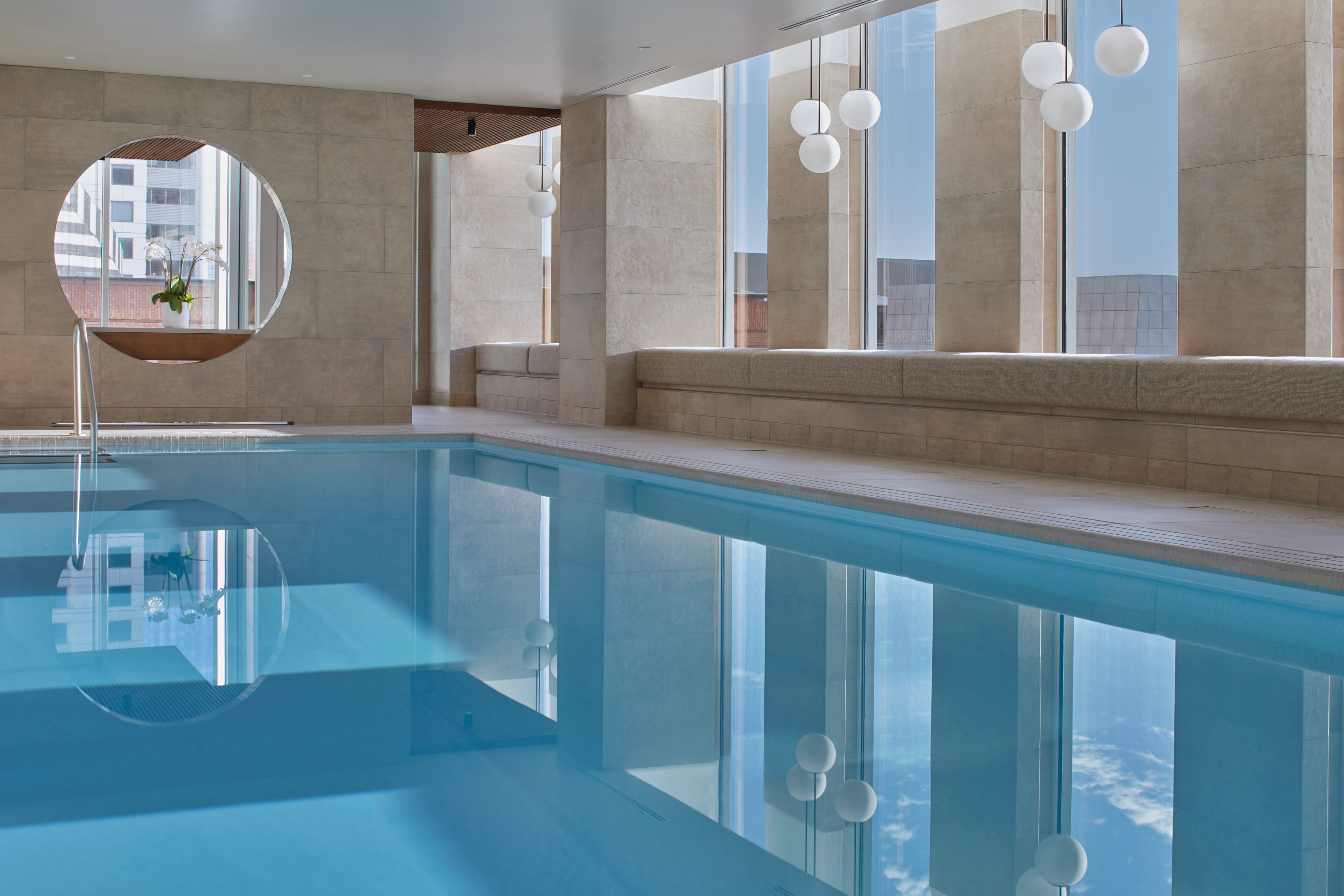 An indoor swimming pool with blue water in a modern room. Upholstered lounge seating lines the pool edge, and circular wall openings reflect in the water. A person walks along the pool deck, and city buildings are visible through windows.