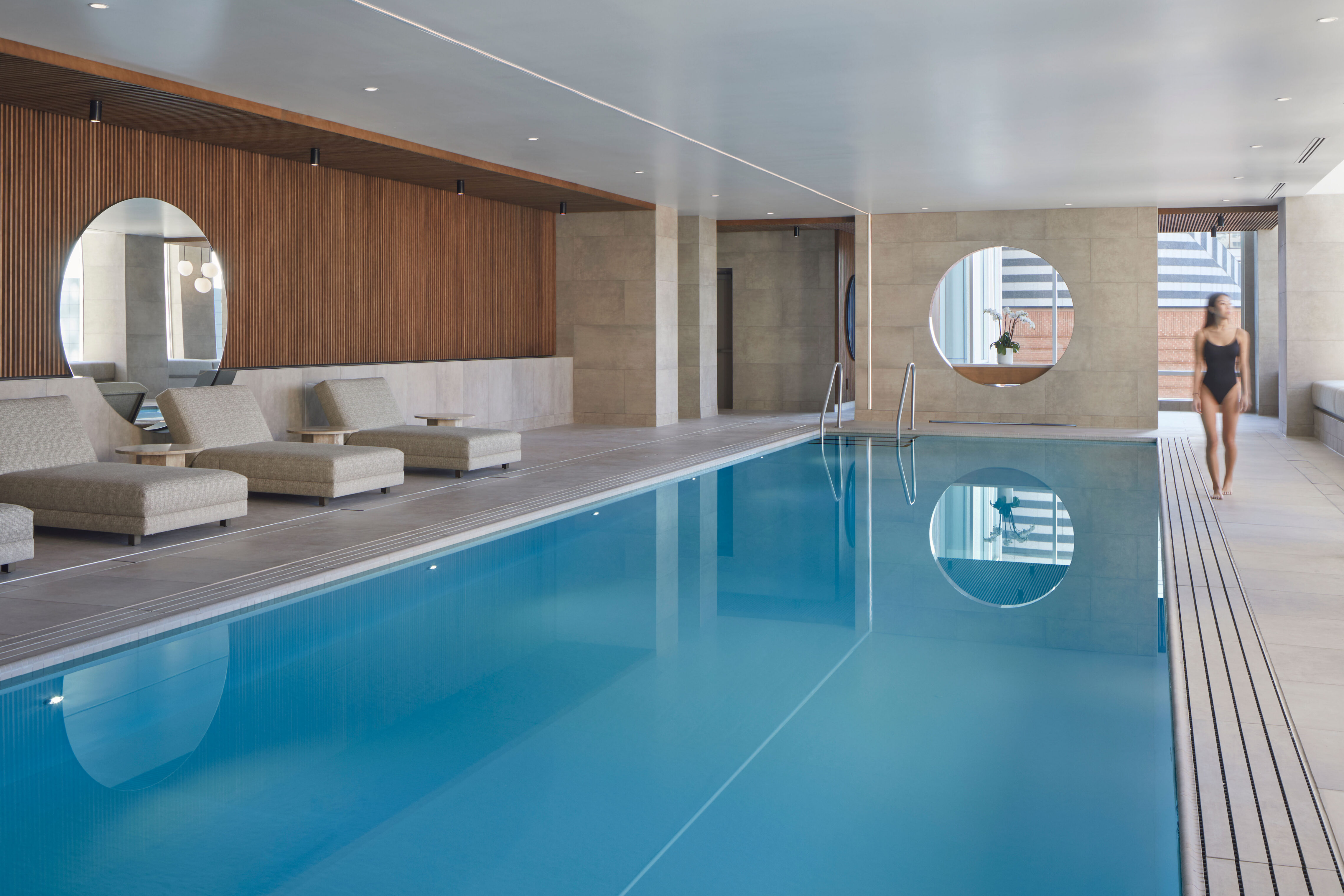 An indoor swimming pool with blue water in a modern room. Upholstered lounge seating lines the pool edge, and circular wall openings reflect in the water. A person walks along the pool deck, and city buildings are visible through windows.