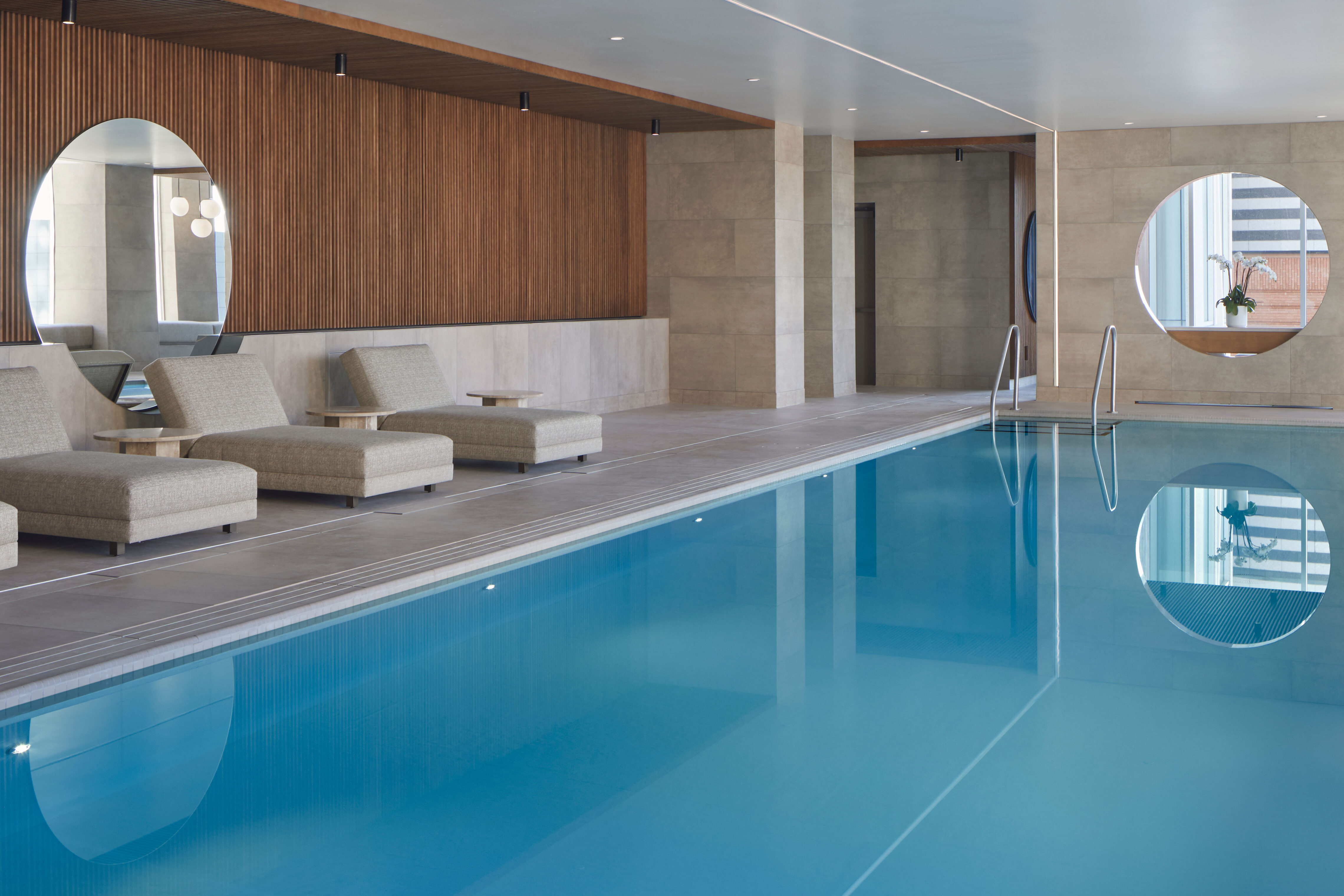 An indoor swimming pool with blue water reflecting circular wall openings. Upholstered lounge chairs and small side tables line the pool deck, with wood slat walls, tiled surfaces, and metal pool handrails visible throughout the space.