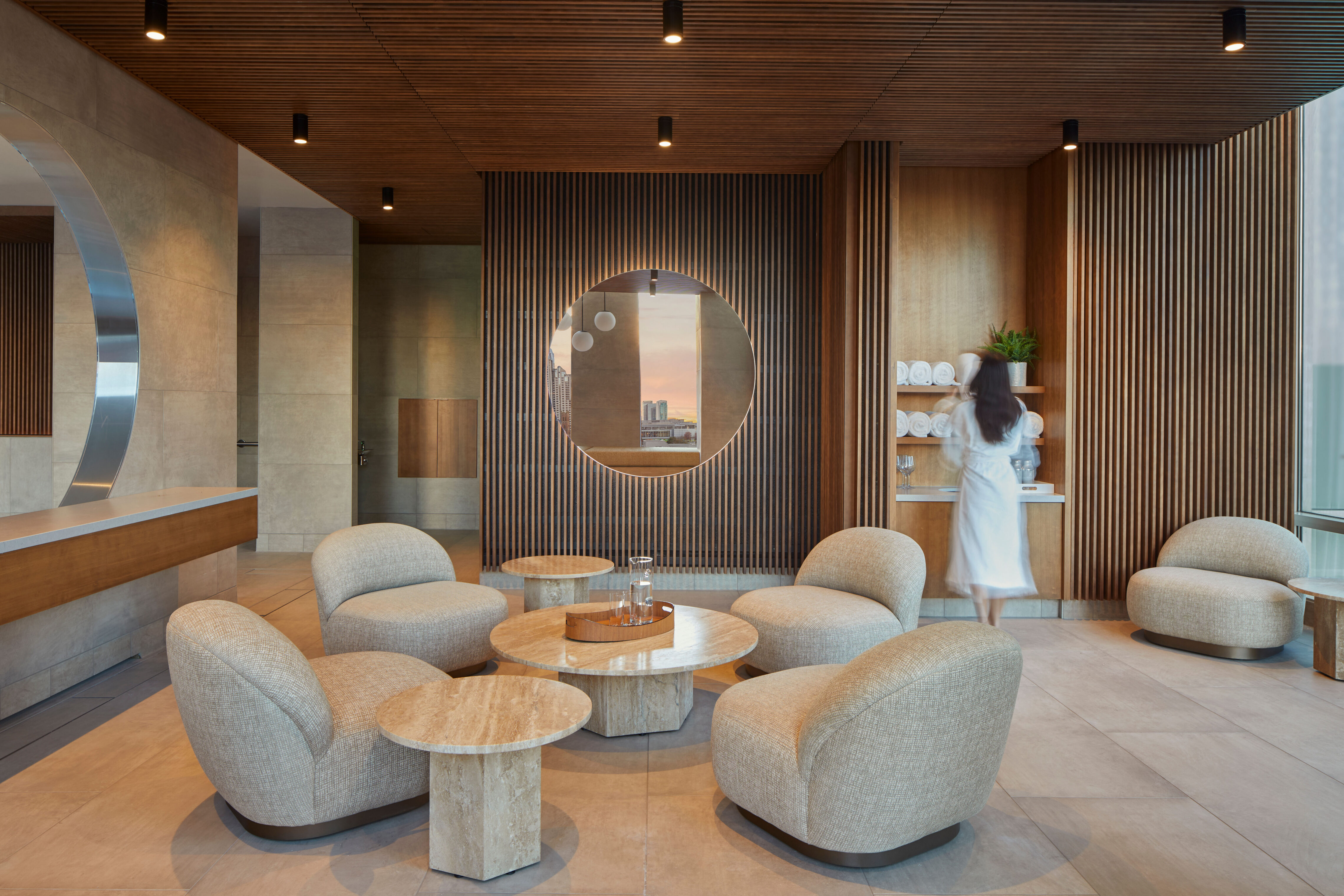 A seating area with upholstered lounge chairs arranged around small round tables in a modern interior. A circular wall opening is centered on a wood slat wall, with shelving holding folded towels and glassware to one side. A person in a robe walks through the space, and ceiling lights illuminate the room.