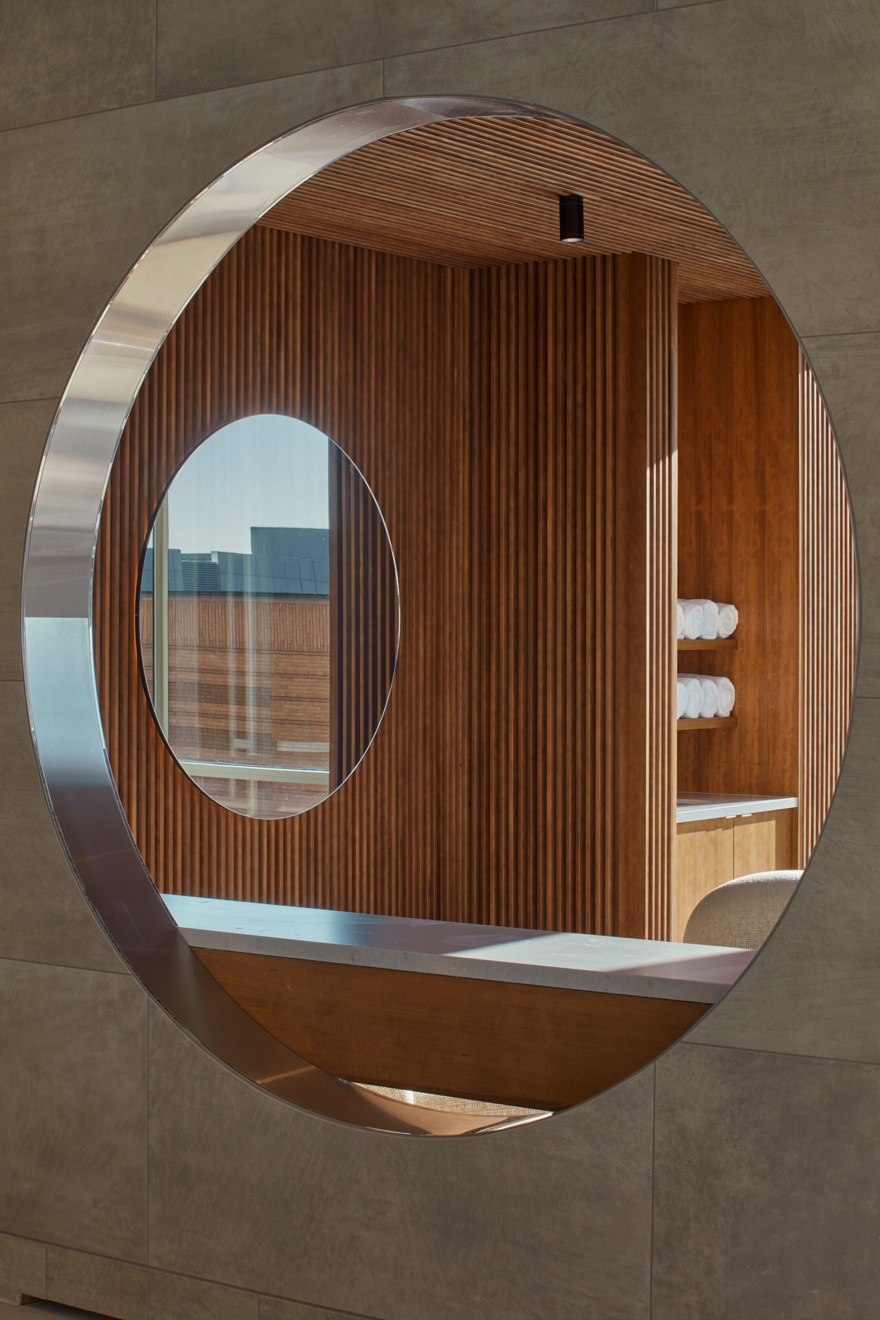 A circular opening framed in metal set into a tiled wall, revealing an interior space with vertical wood slats, built-in shelving with folded towels, and a counter surface. A window and part of a seating area are visible through the opening.