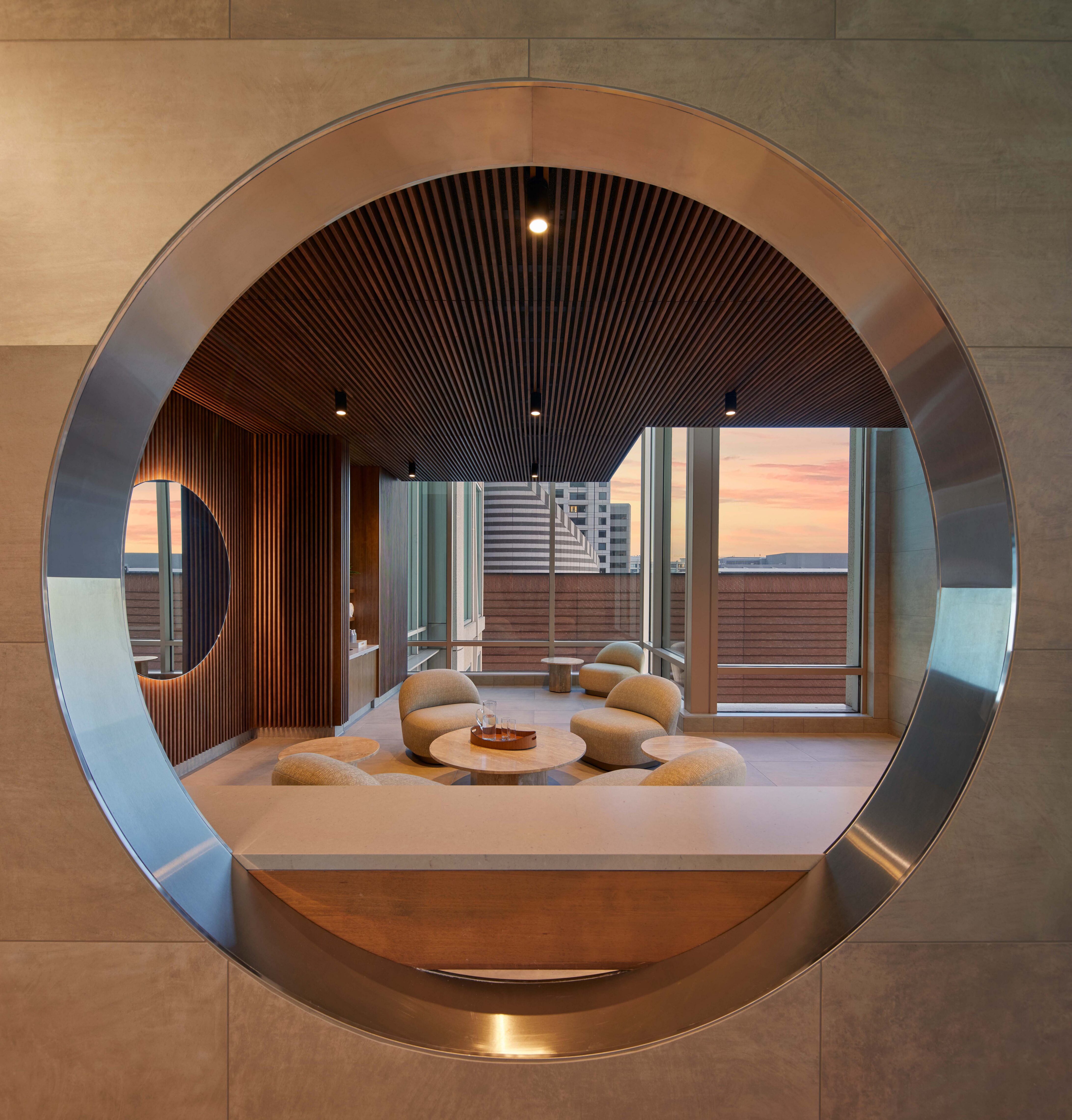 A circular metal-framed opening set into a tiled wall, looking into a seating area with upholstered chairs and small round tables. Large windows reveal city buildings and a sunset sky beyond.
