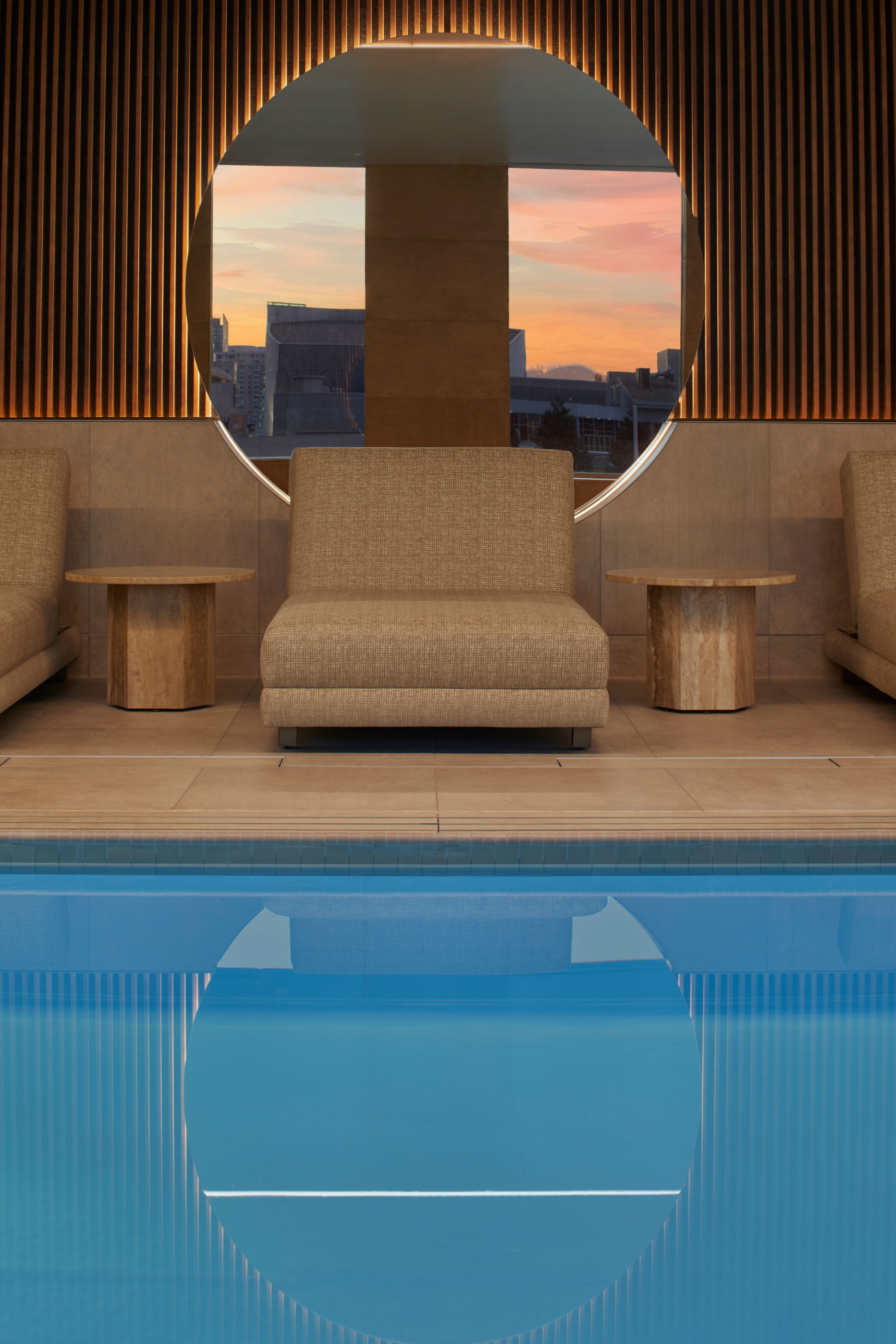 An indoor swimming pool with blue water reflecting a circular wall opening that looks out toward a city skyline. Upholstered lounge seating and small side tables sit along the pool edge, with vertical wood slats and warm lighting on the wall behind.