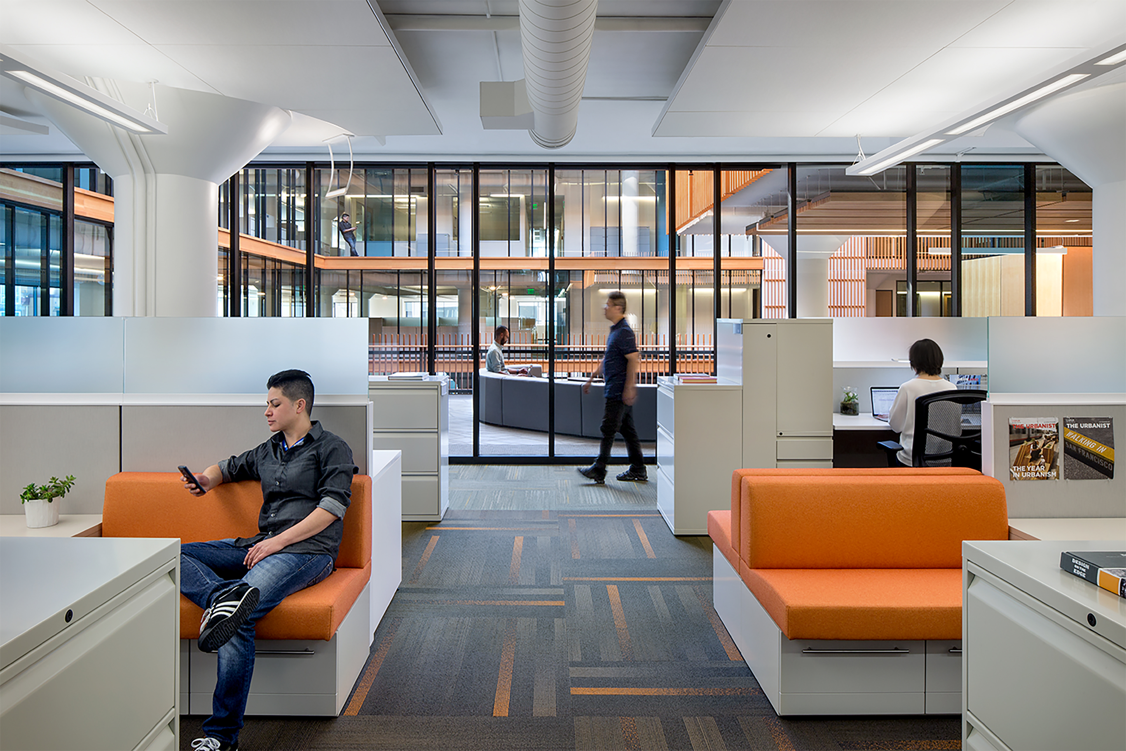 Open-plan office interior with flexible workstations, integrated seating, and glass partitions, showing people working, moving through shared spaces, and using a variety of settings that support collaboration and individual focus.