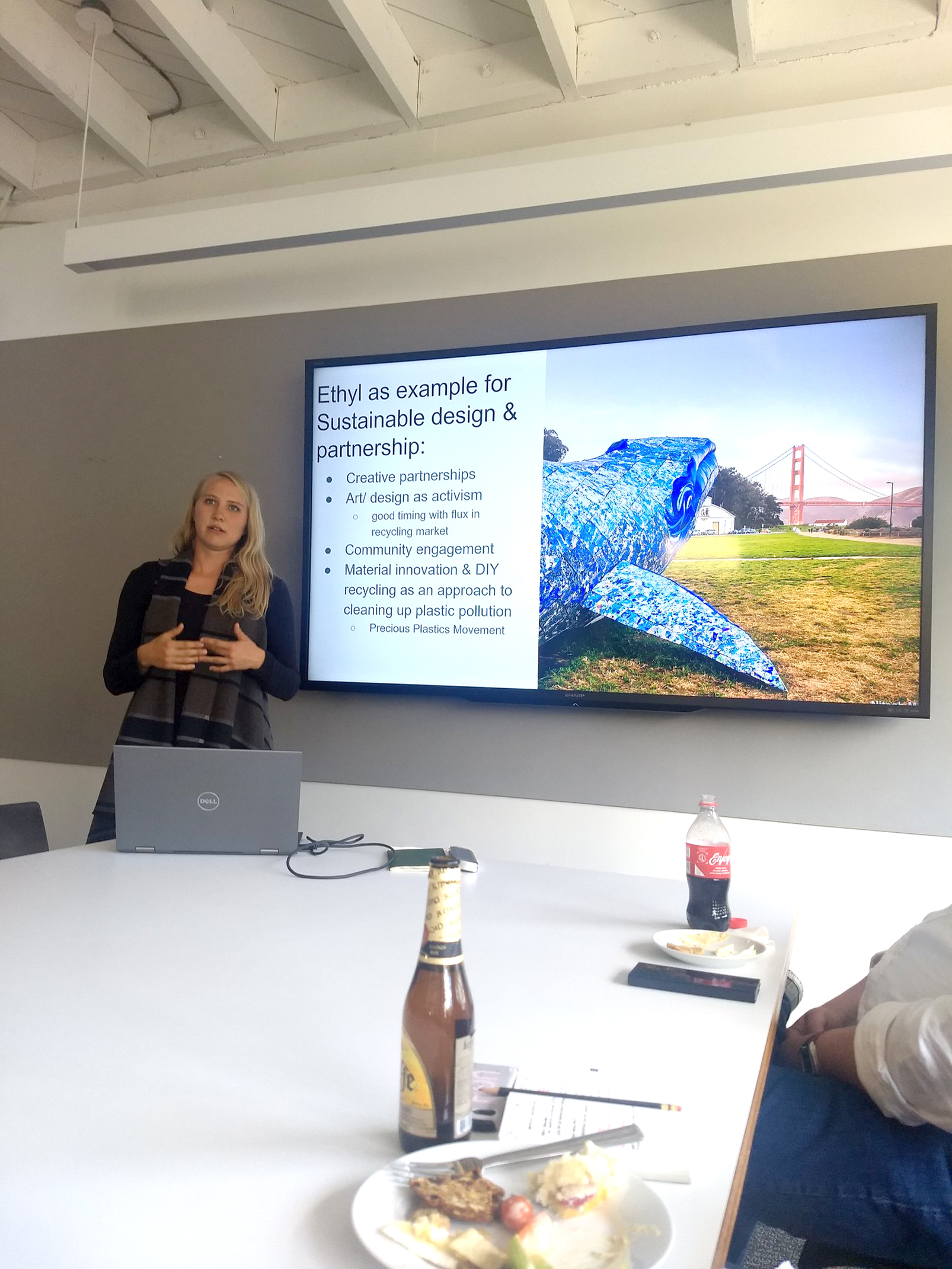 A presenter speaks at the front of a conference room beside a screen displaying a slide on sustainable design and partnership, while colleagues sit around a table with laptops and food during an informal office talk.