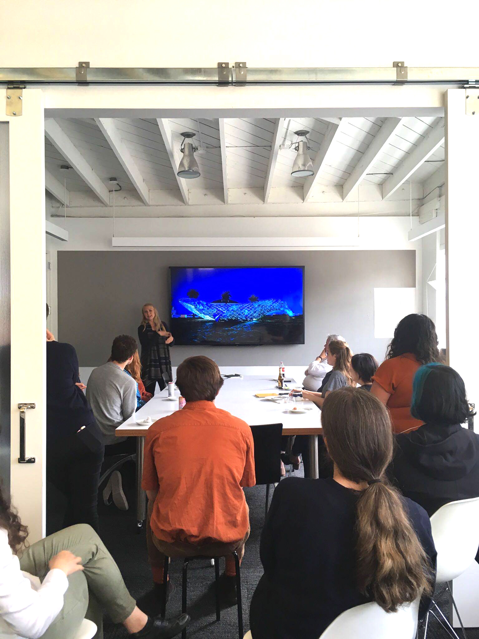 A group of colleagues gathers around a conference table as a presenter speaks and gestures toward a screen displaying a design image during an informal office talk.